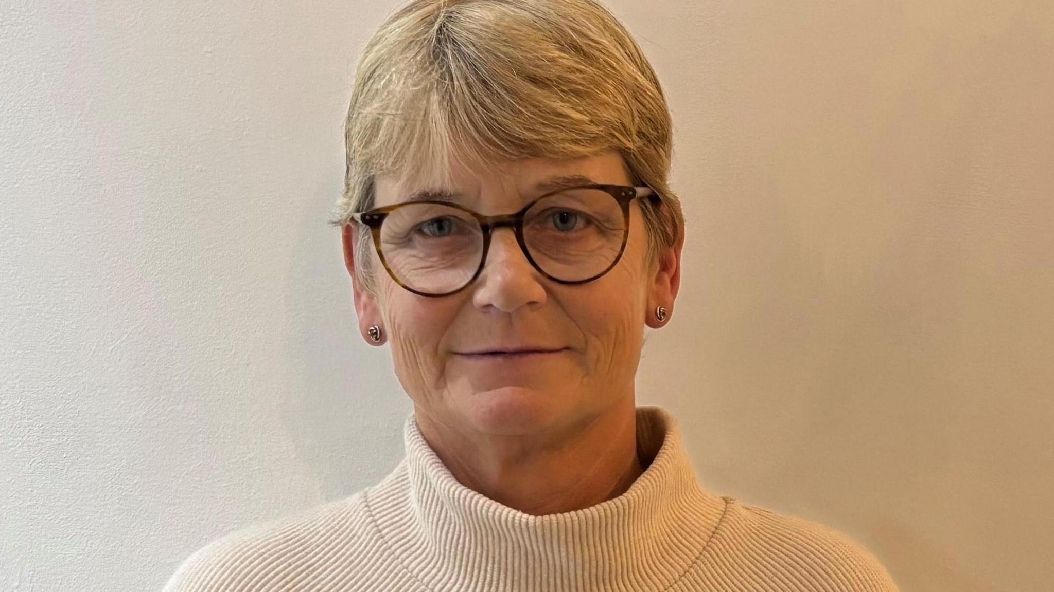 Loraine Wilson (glasses, short blonde hair) looking straight to camera. Wearing a cream top.