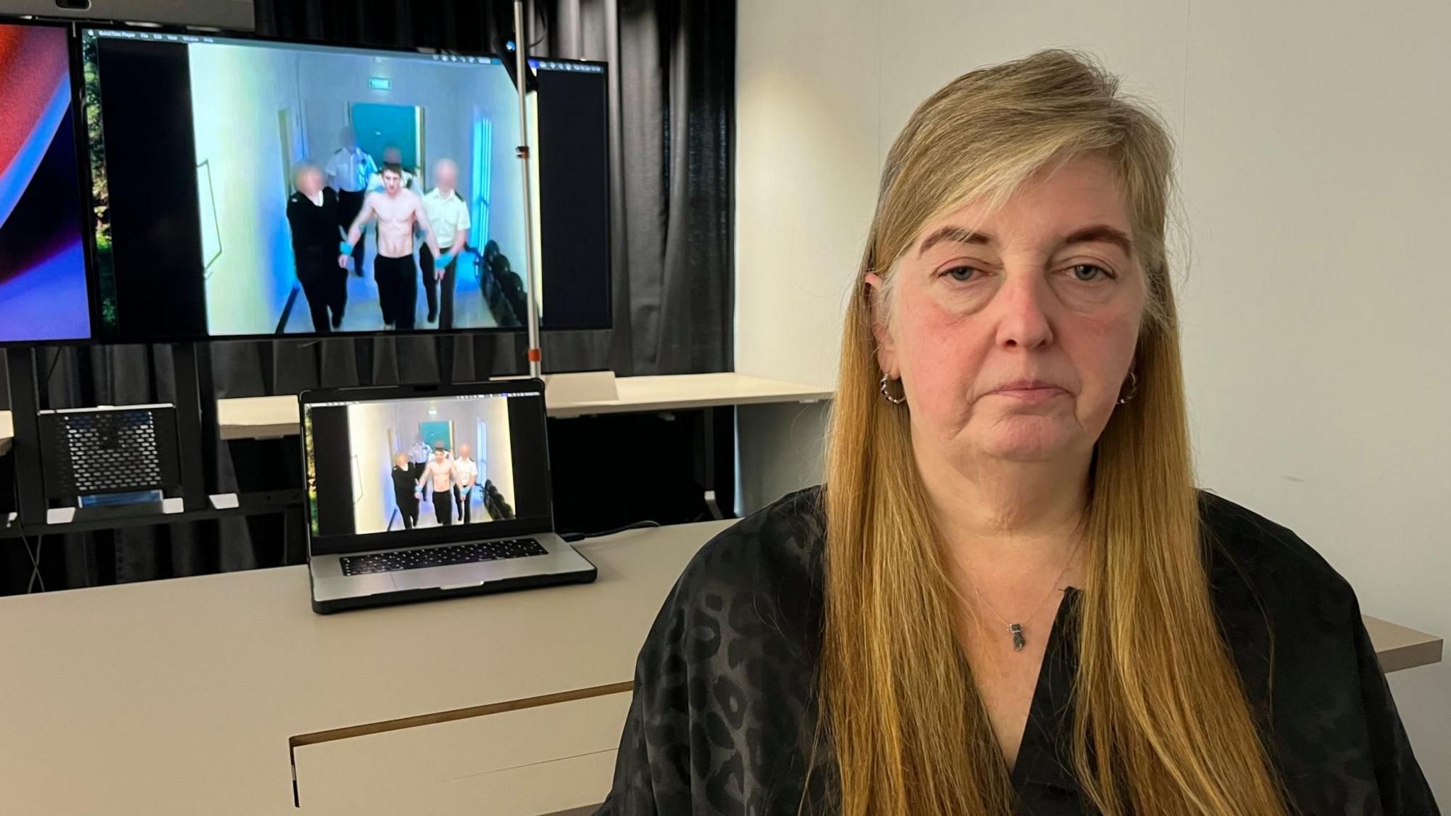 Sharon McFadyen seated indoors in front of a desk, with multiple screens behind showing a video of a shirtless individual being escorted down a corridor by several others; a laptop on the desk mirrors the same footage, and the room includes a white wall, shelving, and dark curtains.