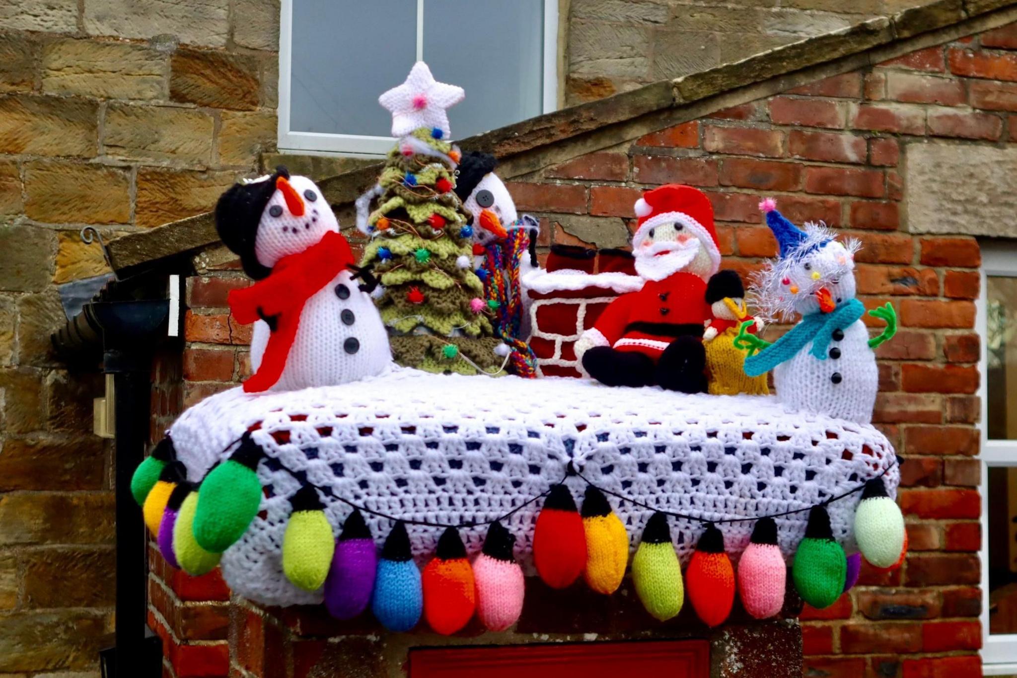 A knitted scene depicts a three snowmen and Santa gathered around a Christmas tree.