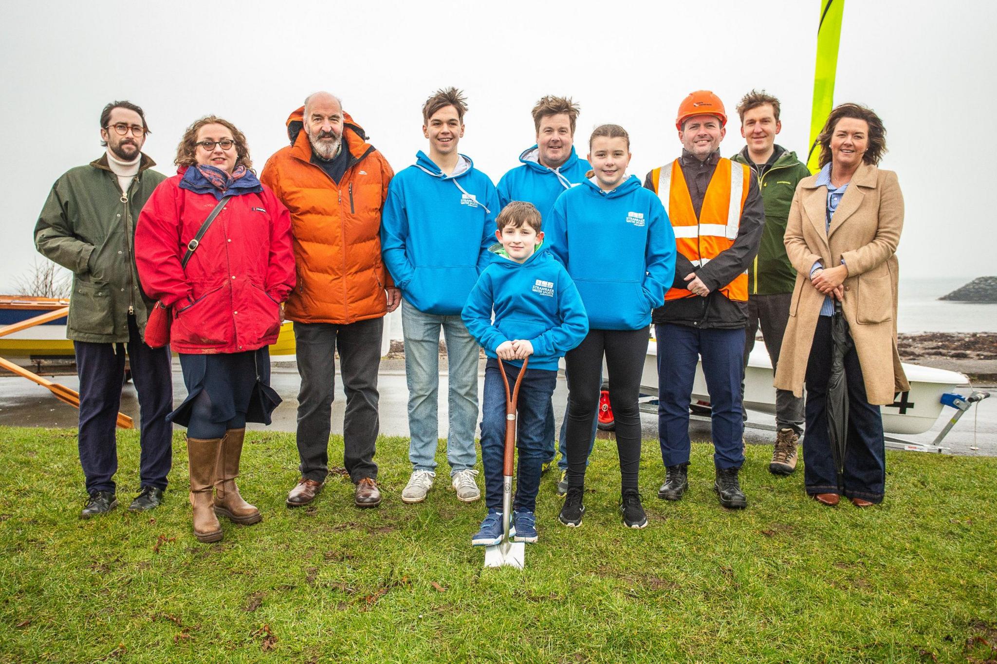 A group of people line up behind a young boy with a spade cutting the turf on a new water sports centre for Stranraer