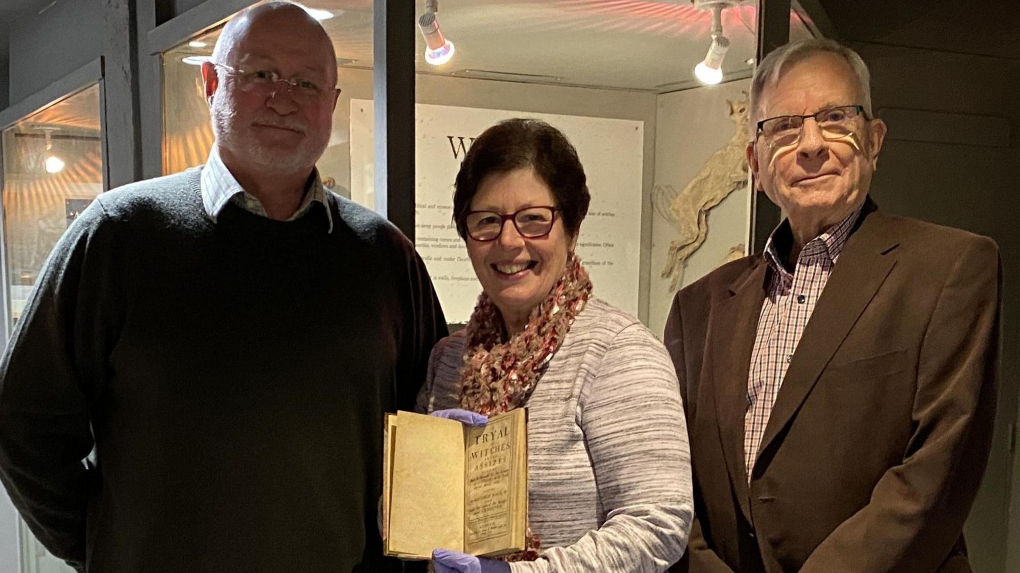 Two men stand alongside a woman who holds the book while she wears blue gloves. They smile at the camera.