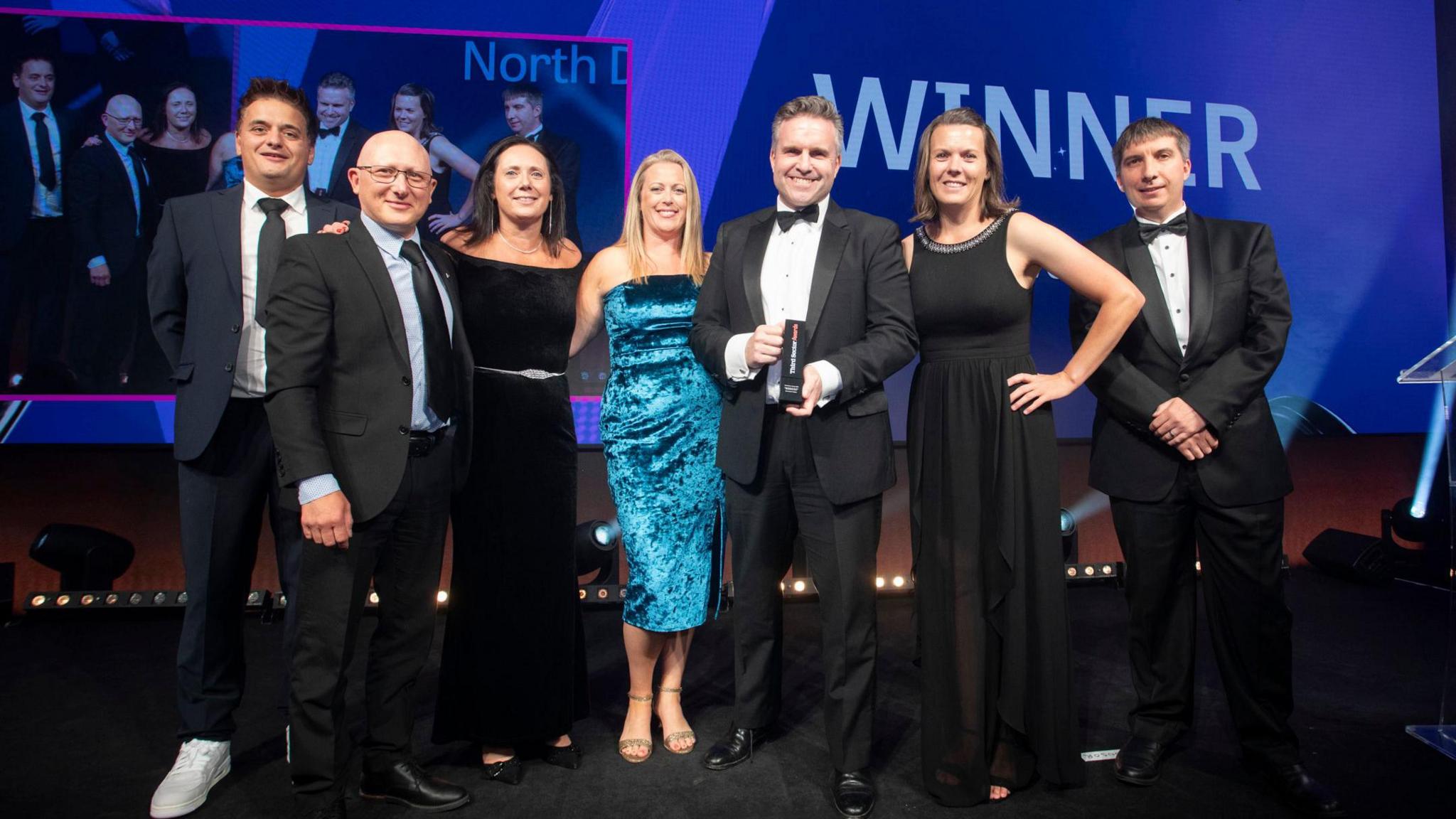 A group of people smartly dressed at the awards. A man wearing a tux is holding the area. The group, made up of men and women, are standing in a row and are smiling at the camera. A large purple board in the background says winner.