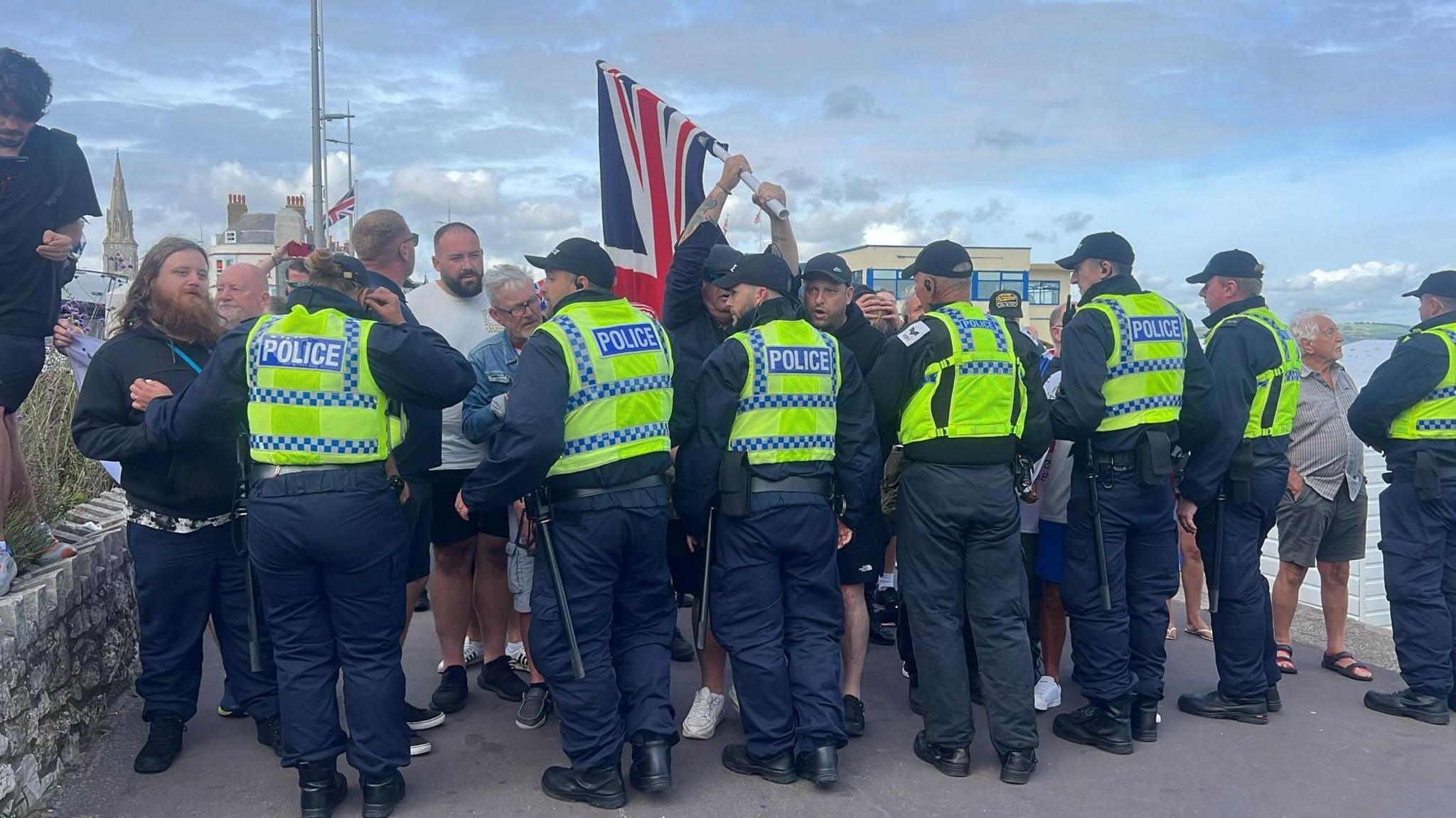 Seven police officers stand in a line in front of a group of men, one of whom is holding a Union Jack.