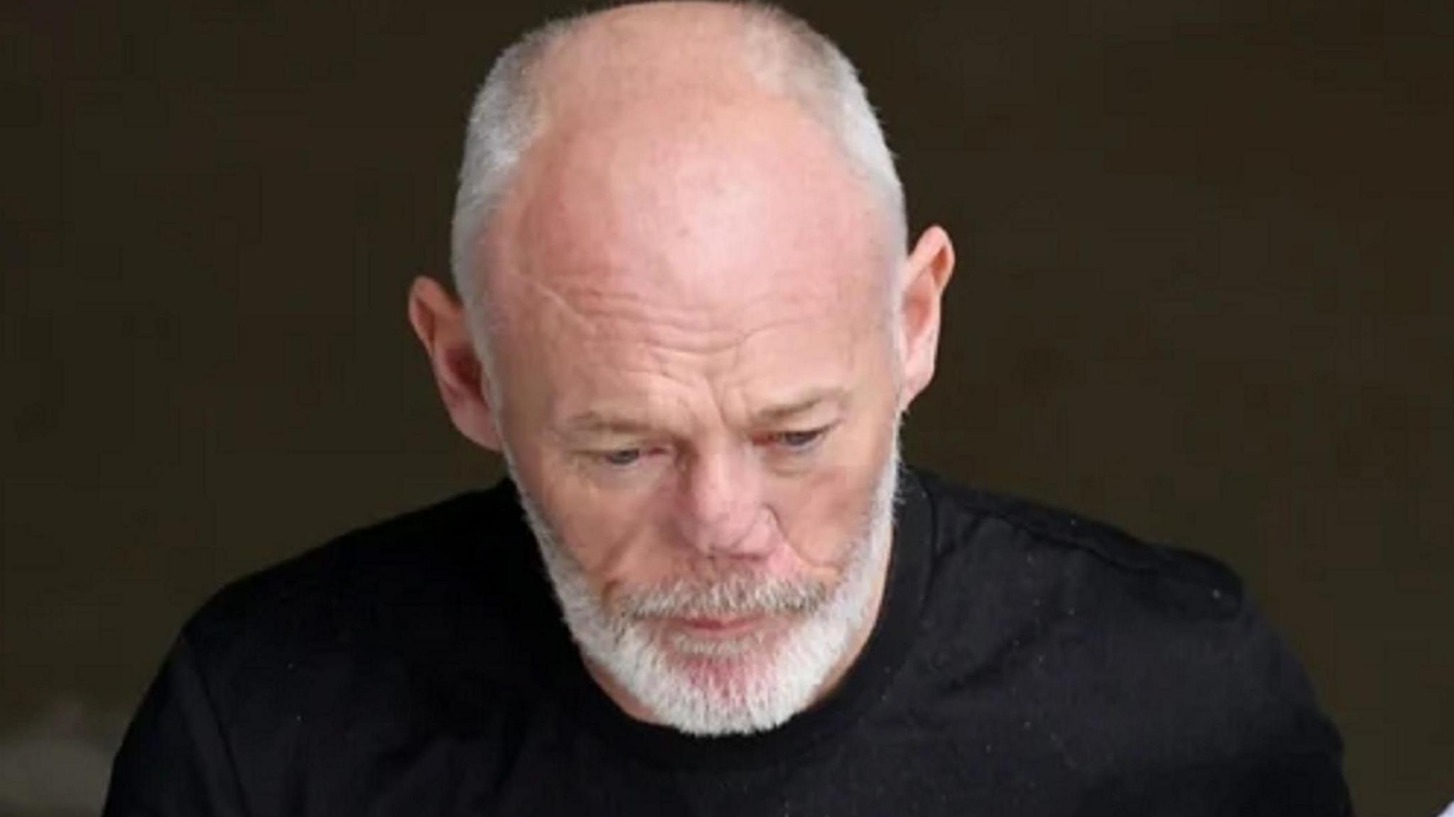 Andrew Pender is looking towards the ground, he is semi-bald with short grey hair and a great beard and moustache. He is wearing a black tee-shirt