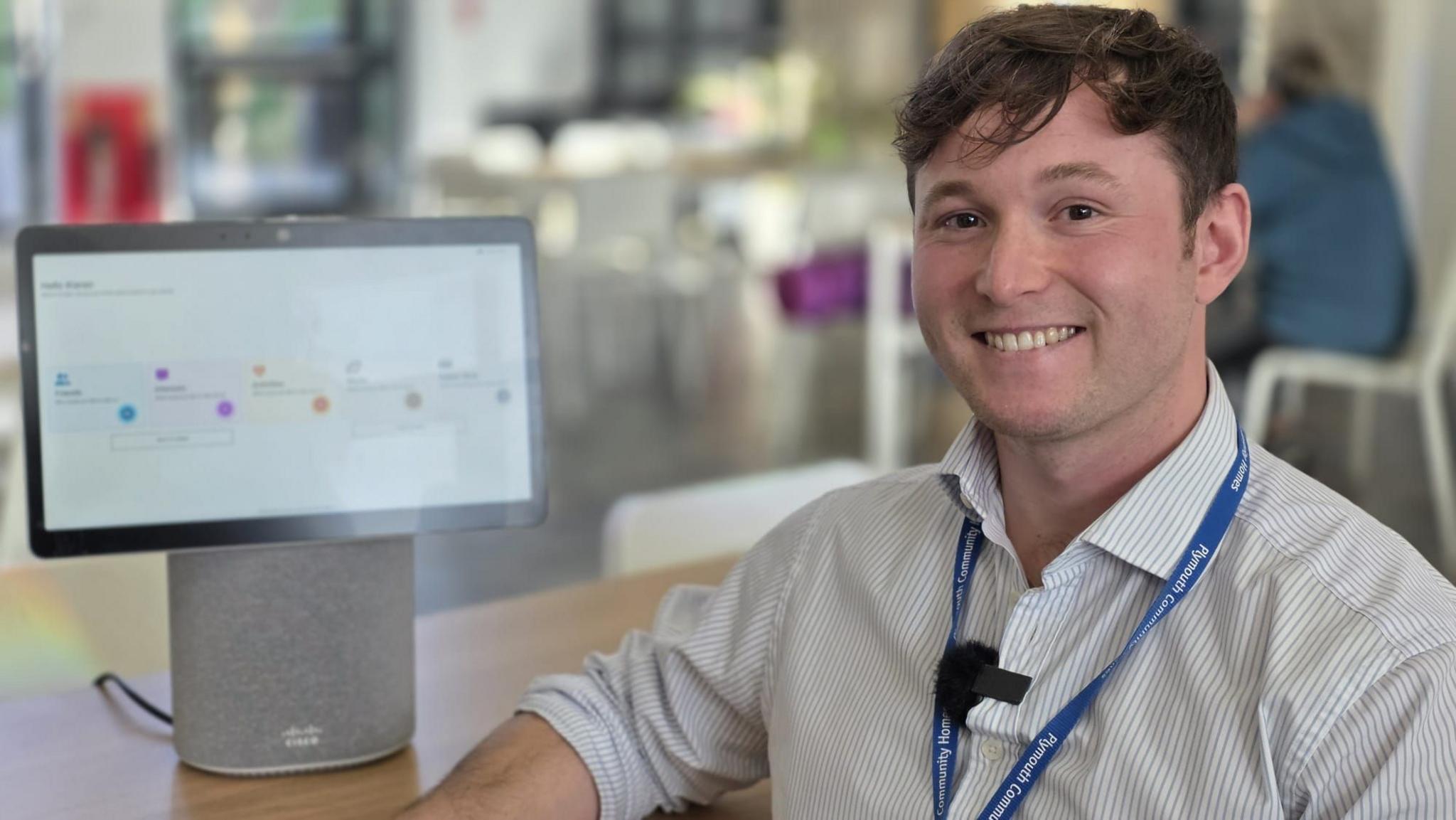 A man with short brown hear, wearing a white striped shirt and smiling, with a computer screen behind him