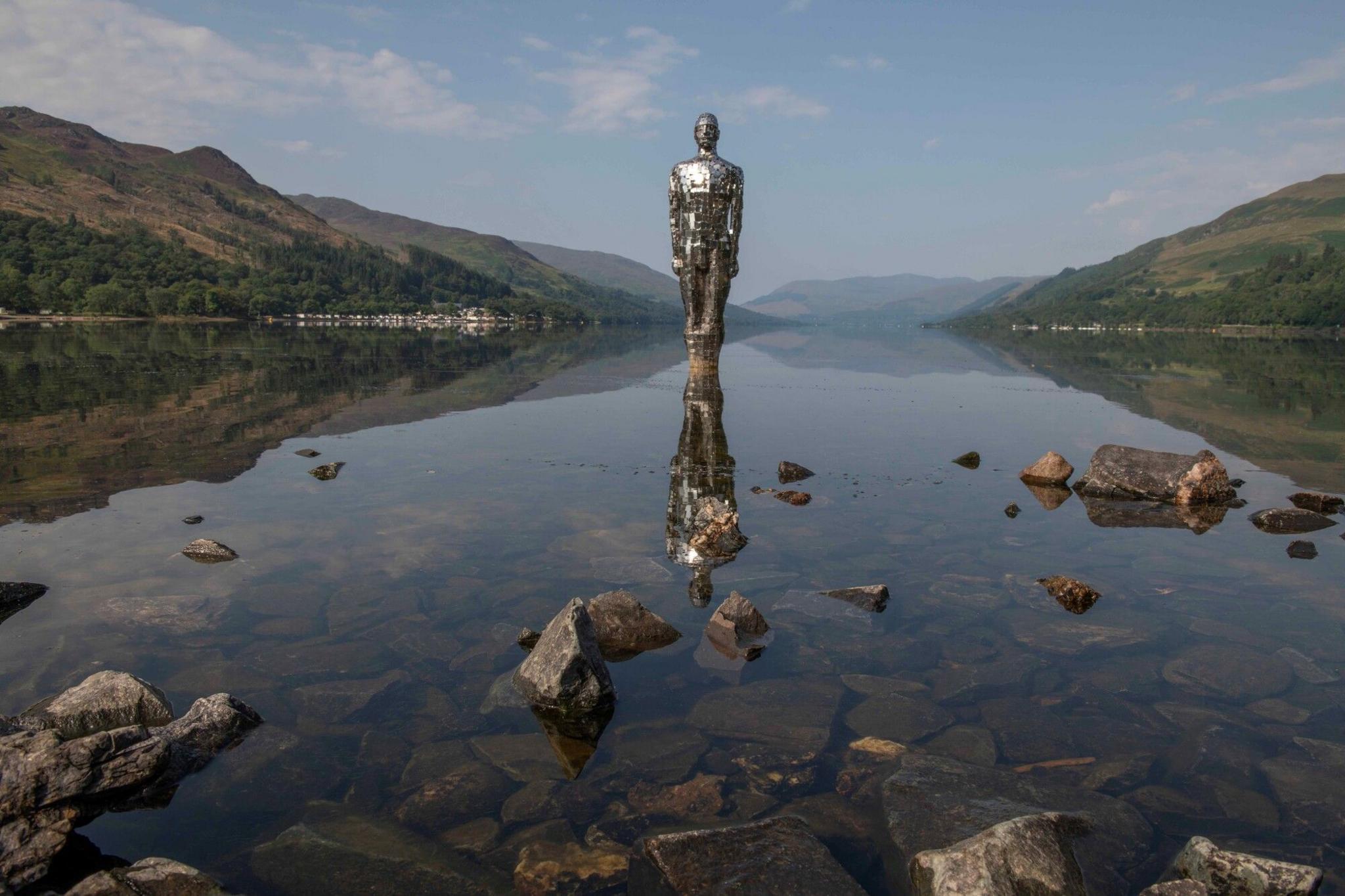 Mirrored statue of a man standing in a still loch with some large rocks poking out towards the shore and surrounded by grassy hills