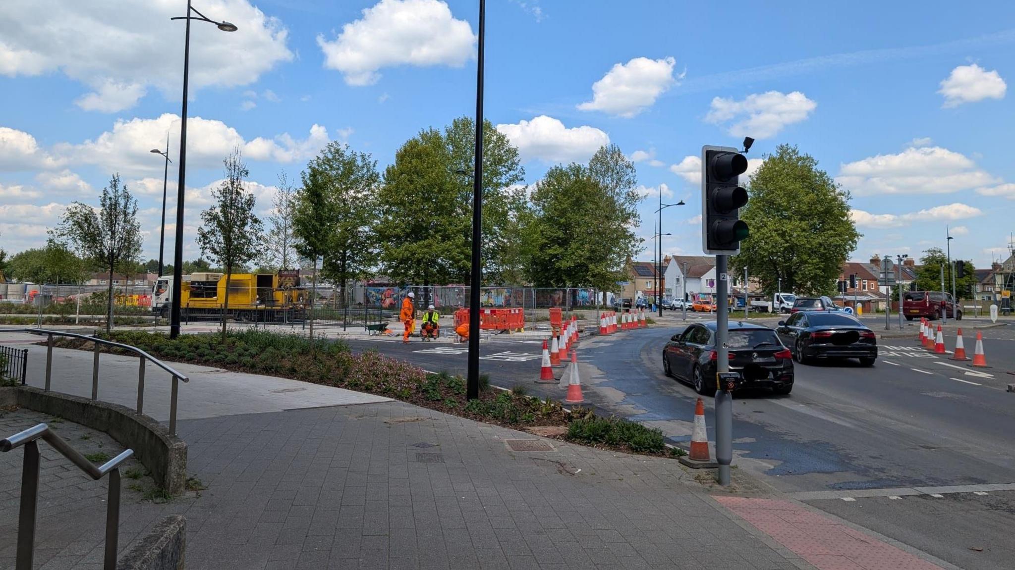 A foot path next to a road where road works are happening. There are a few workers in high vis at the site and two cars are driving past.