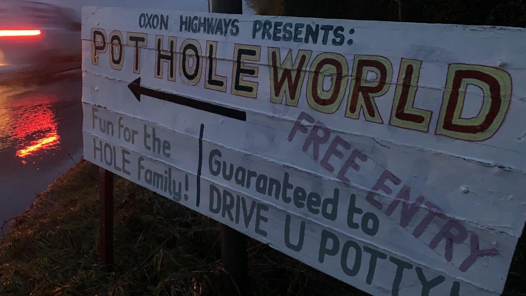 A sign reading: "Oxon Highways presents: Pothole World", "fun for the hole family" and "guaranteed to drive you potty". It is right next to a road.
