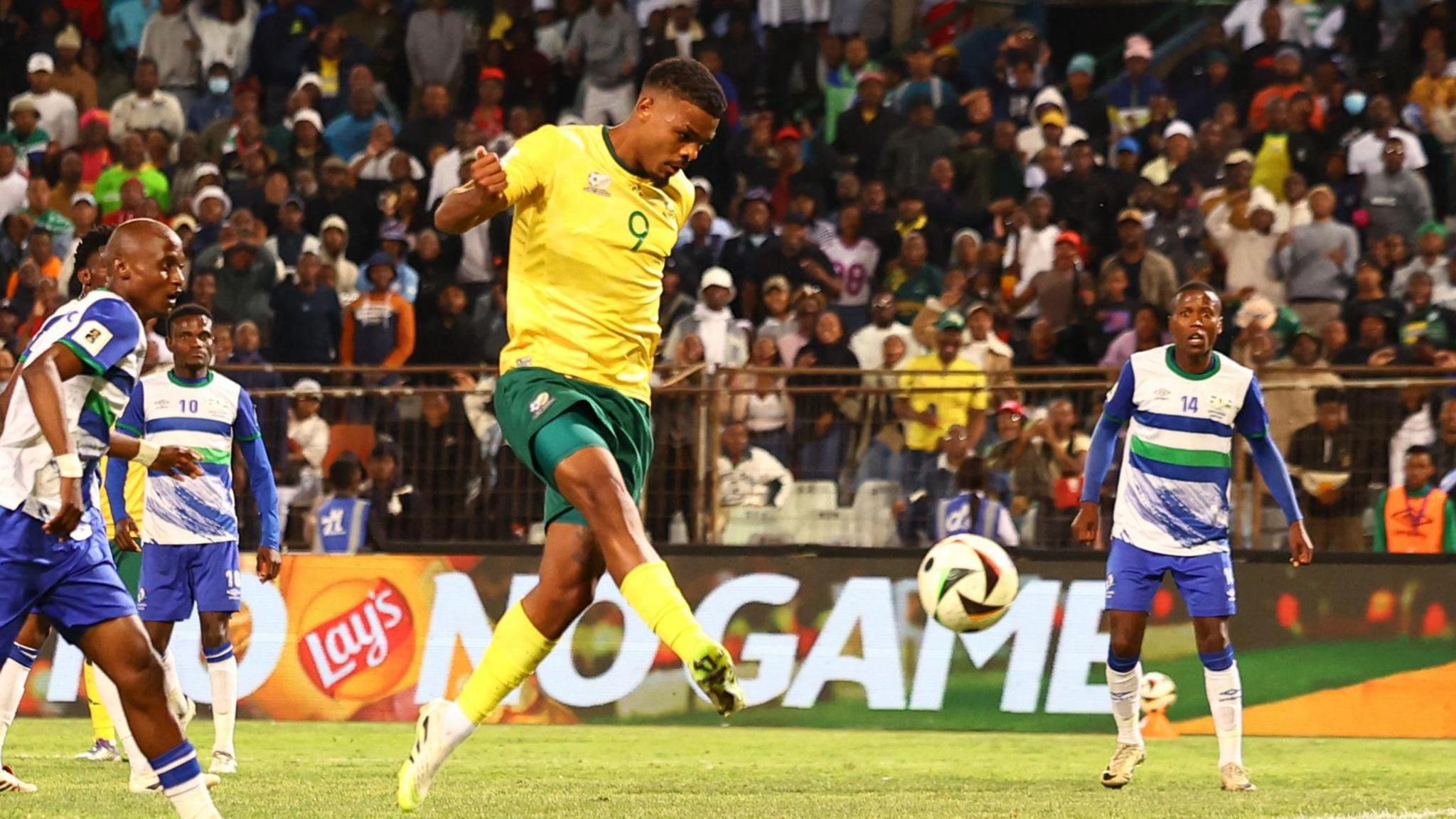 Lyle Foster, wearing a yellow and green South Africa kit, volleys the ball as three Lesotho players watch on and a large crowd is seen in the background