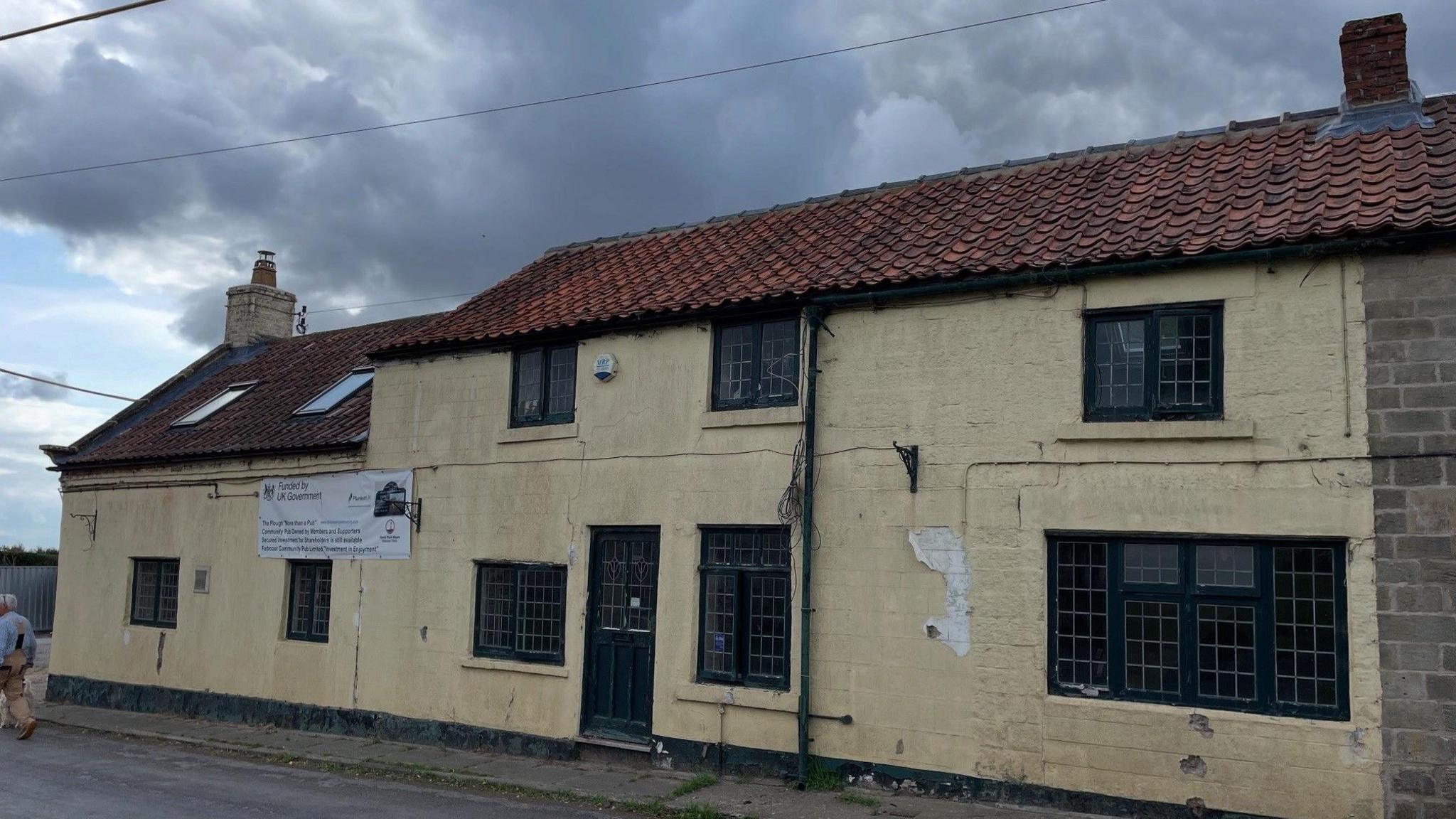 An external shot of The Plough, Fadmoor. A beige sand-coloured pub building with black framed windows, a black door and red tiled roof. There's a white sign affixed to the building labelling it as an Asset of Community Value.