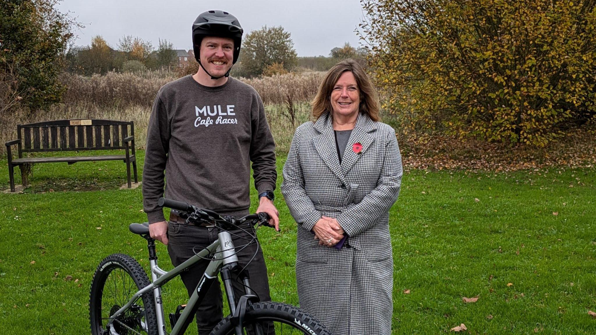 A man and woman stand next to each other on grass in front of a wooden bench. The man is holding up a black bike, wearing a black bike helmet, dark grey jumper and black jeans. The woman has shoulder-length brown hair and is wearing a long black and white checked coat with a poppy at the lapel. Long grass and shrubs can be seen in the background.