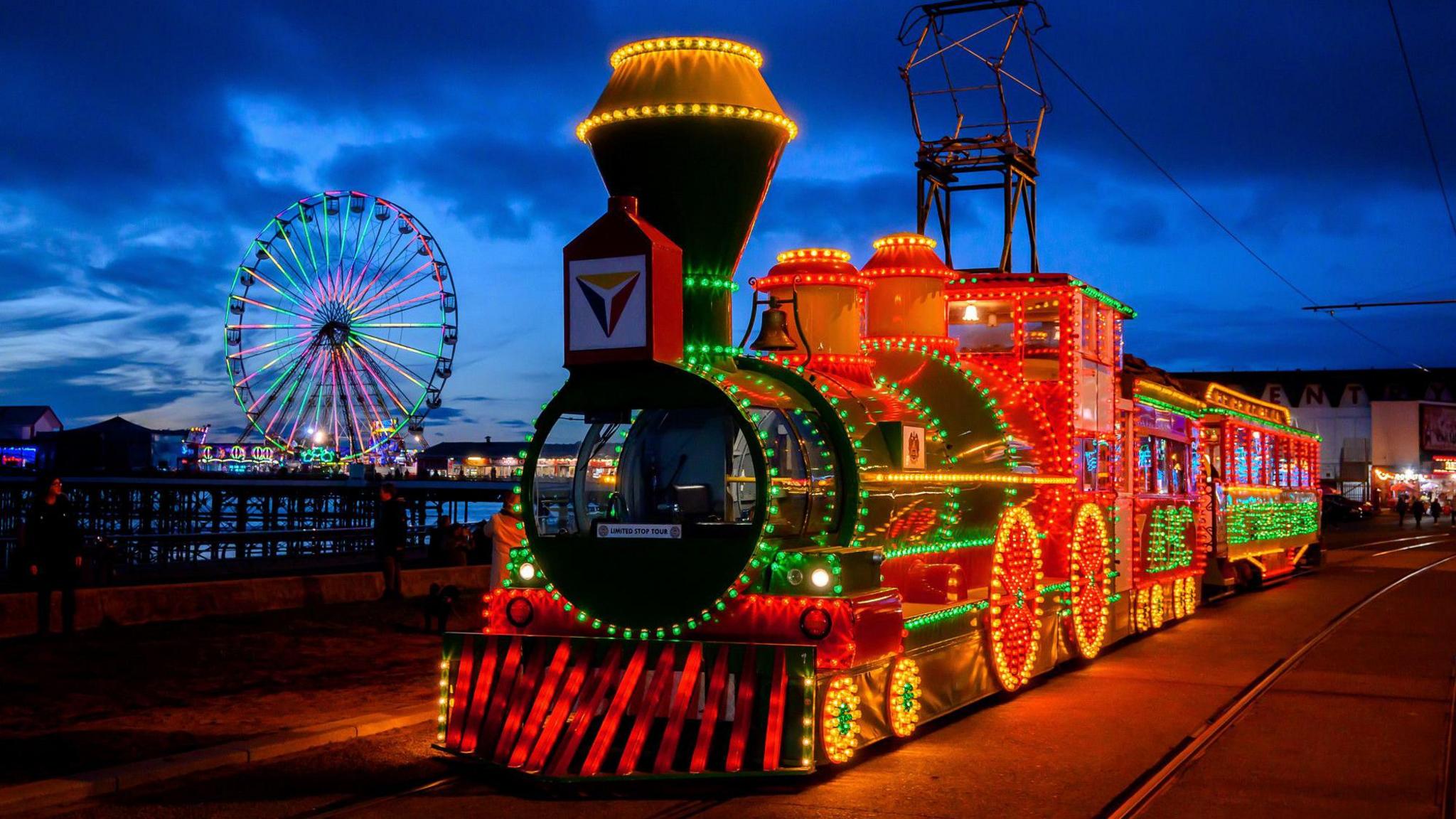 An old tram is covered in red, green and yellow lights travels along Blackpool Promenade at night.