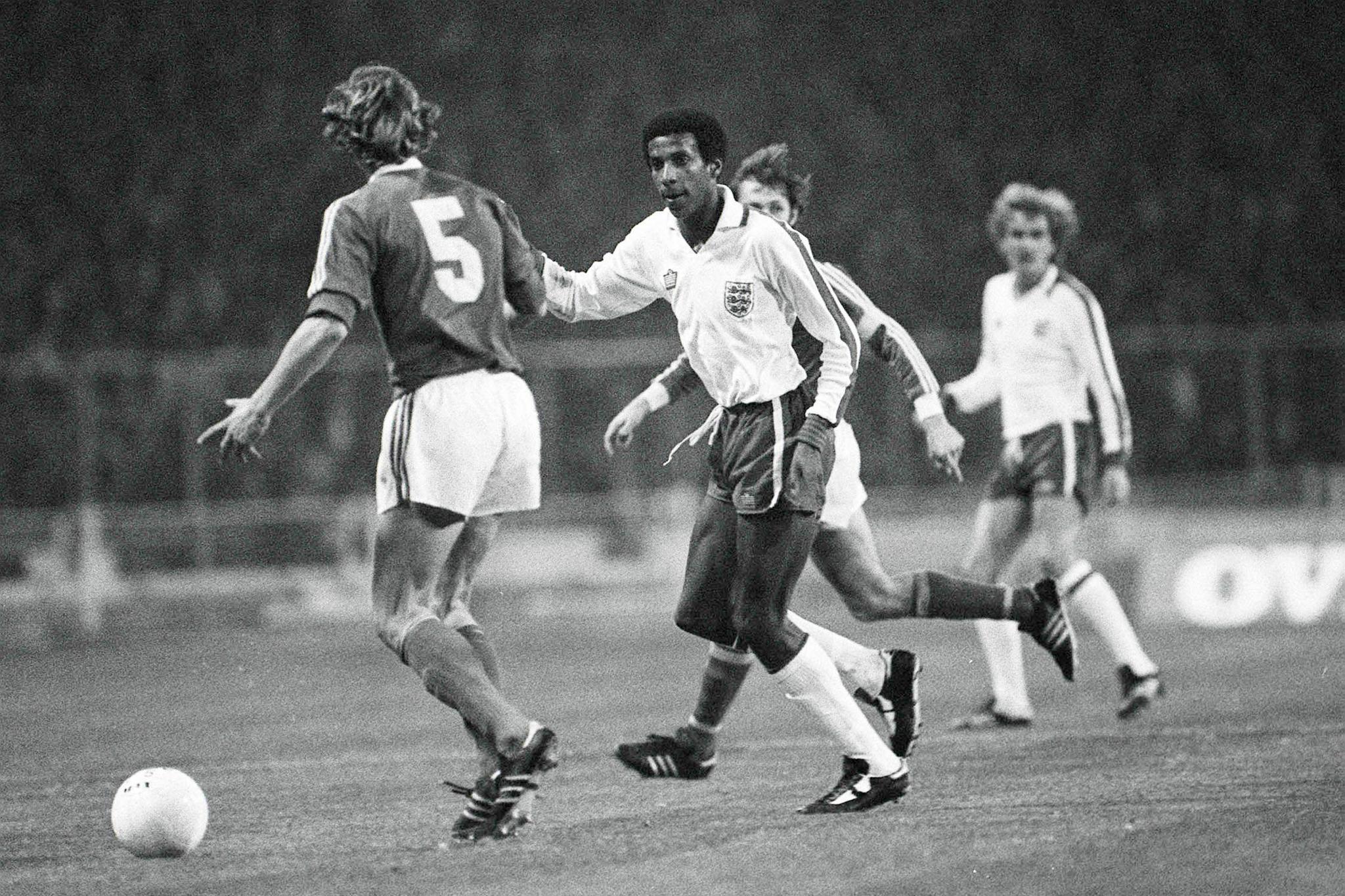 A black and white image of Viv Anderson, wearing a white long-sleeved shirt, dark shorts and white socks, during his England debut. A Czechoslovakia player wears a dark shirt bearing the number 5. His shorts are white.