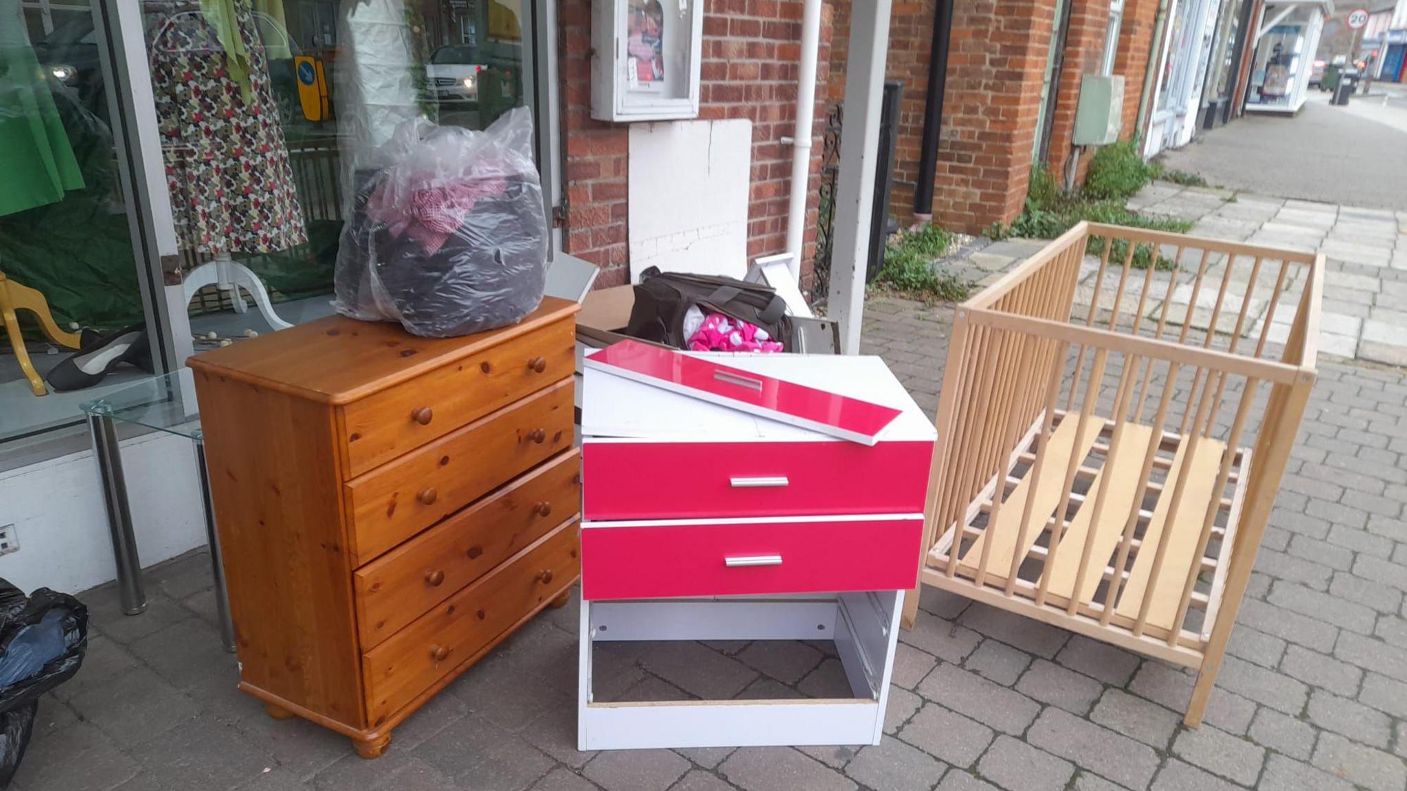 Two sets of drawers, one wooden and one white and pink, have been left outside the front of a charity shop. A wooden crib, missing the bottom, has also been dumped. 