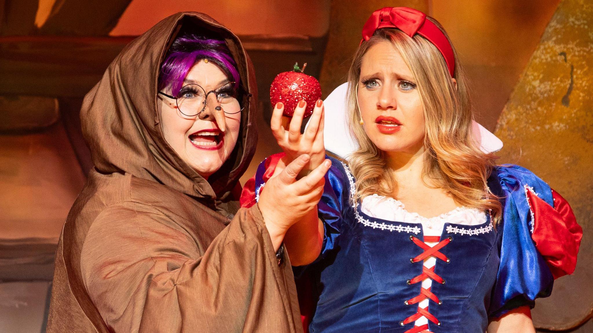 Emma Norman as Queen Grimelda and Olivia Birchenough as Snow White in Snow White and the Seven Dwarfs at Theatre Royal Bath. Emma is dressed in an old hag costume, with a brown cloak and a long nose with a wart. Her mouth is open and she is holding Olivia's wrist. Olivia is holding a bright red apple and has a scared expression. She is wearing a blue and white costume and a red headband. 