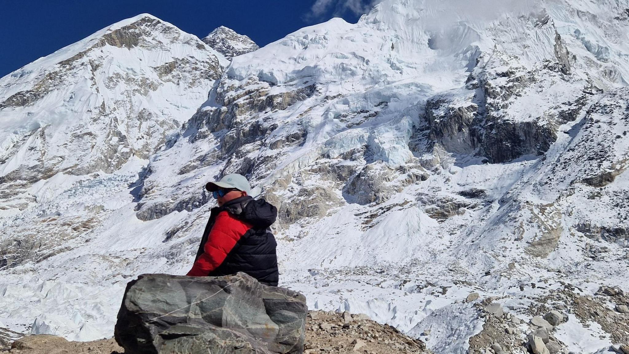 Fergus Maclean is wearing sunglasses, a cap and red and black puffed jacket. He is sitting next a boulder with high, jagged snow-covered mountains behind him.