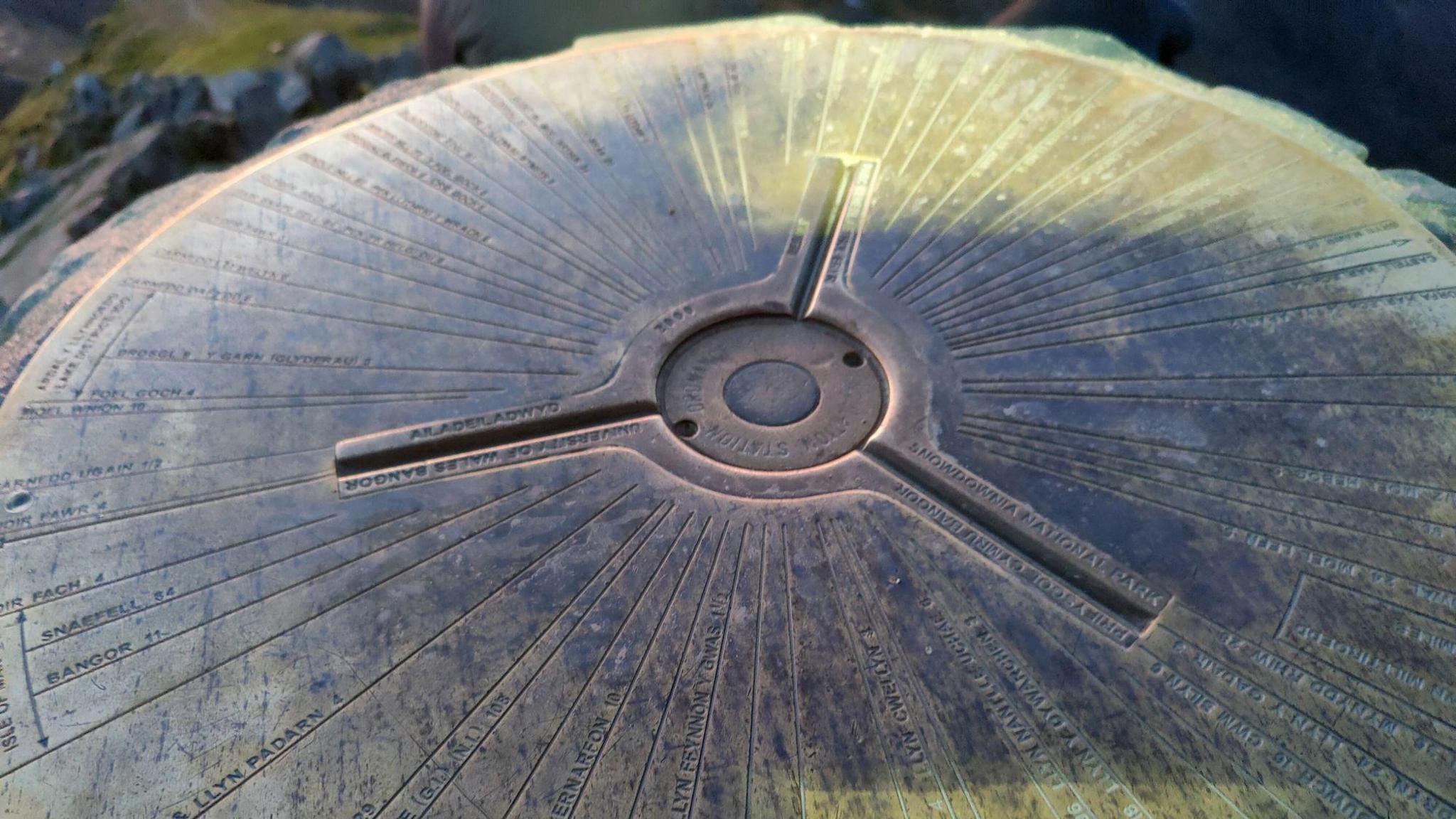 A metal plate can be seen on top of the trig pillar. There is a small circle with three legs coming out from it. 