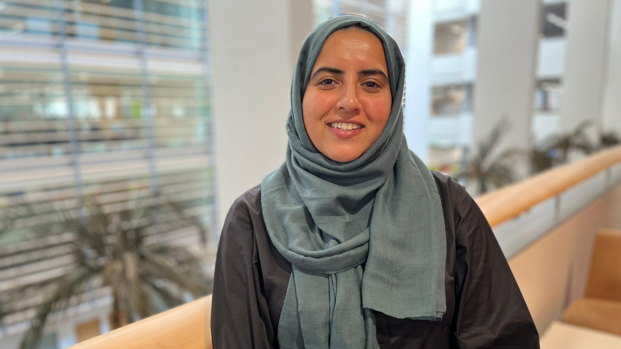 A woman wearing a headscarf in a dark green colour. She is wearing a dark coloured top.