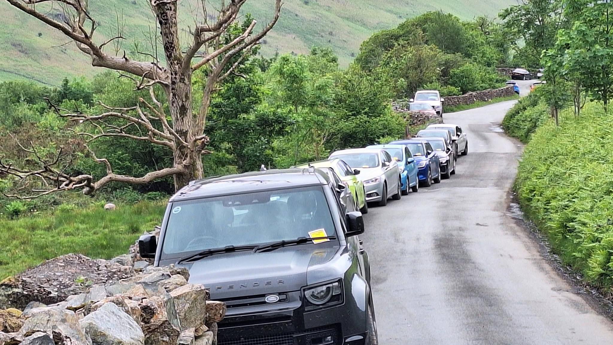 A row of vehicles parked along a narrow country road. The Land Rover at the front of the line has a yellow parking ticket stuck to its front window. Fells are to the left, with trees lining the road.