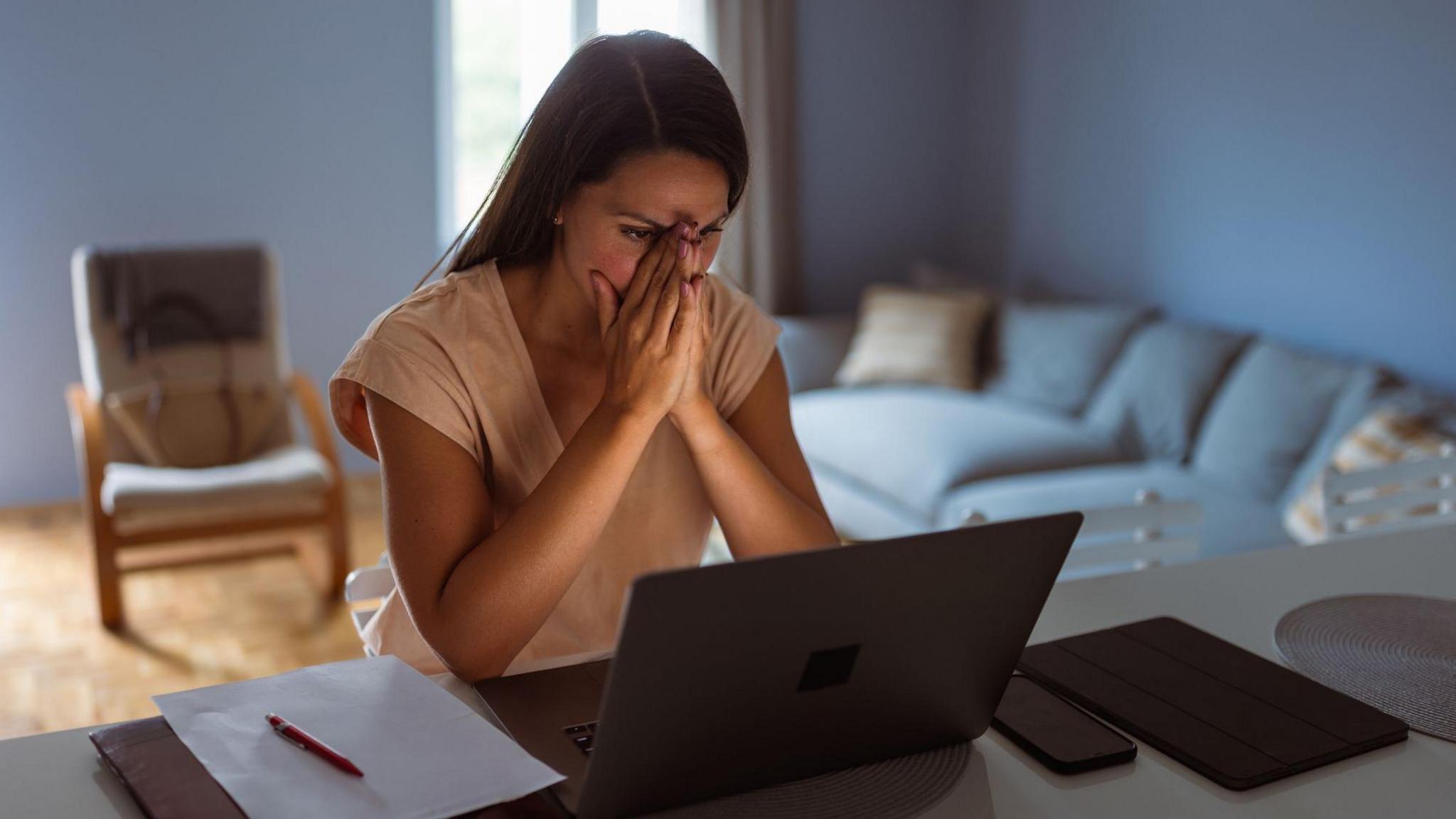 Young woman with her face in her hands looking distressed at a laptop screen with her mobile phone, ipad and paper and pen next to her