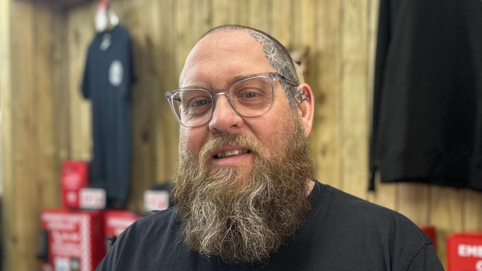 Jamie Hart smiles at the camera. He is bald, has a tattoo on one side of his face and has a long light coloured beard. He wears a black T-shirt.