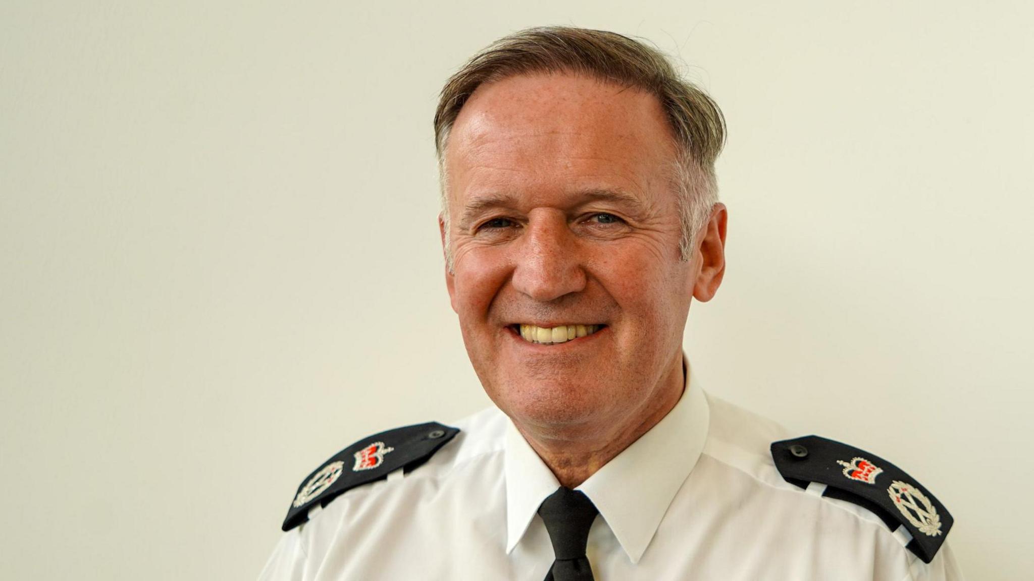 A head-and-shoulders picture of Temporary Chief Constable Darren Martland. He is wearing a white police shirt with black tie. He has short salt-and-pepper hair combed over with a side parting. He has blue eyes and he is smiling at the camera.