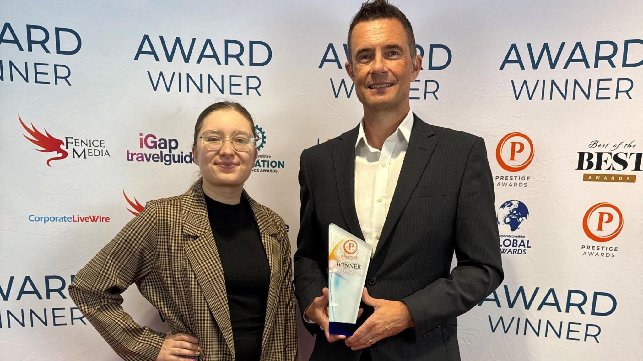 A young woman in a gingham-style blazer and glasses stands next to a man in a dark suit and white shirt. He is holding a glass award which says "winner" on it.