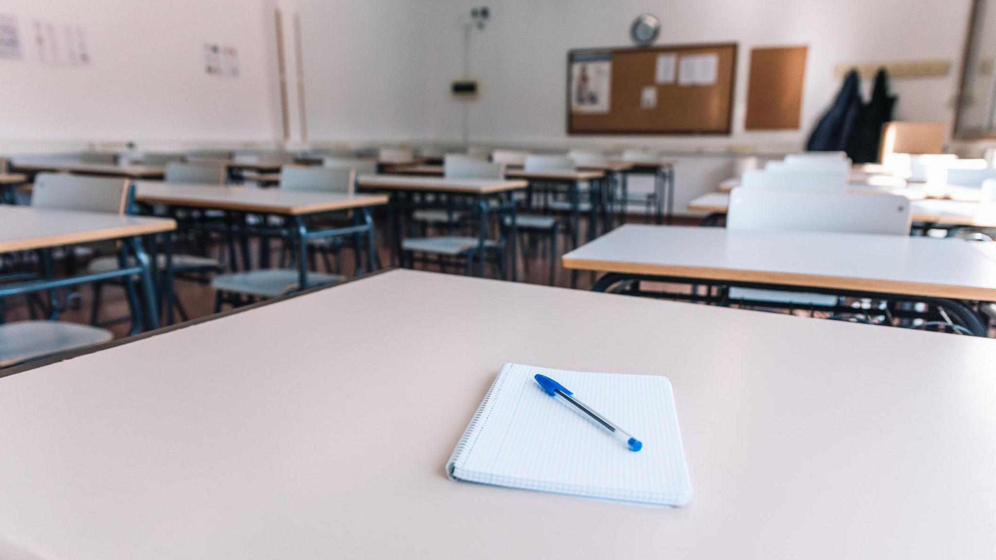 Stock image of a classroom. There are a number of grey desks. The one in the foreground has a notepad and pen on the top.