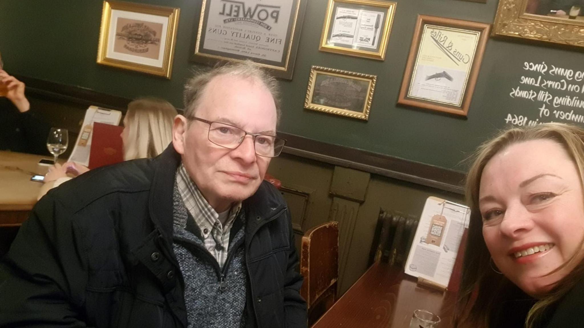 A man looks into the camera while sat at a dark wooden table. He has short greying hair and wears rectangular, thin-rimmed glasses. He has a grey, thin checked shirt on, with a darker grey jumper over that and a black coat over that. Just visible to his right is a woman with light brown hair, also looking into the camera. Behind them on a dark green wall are several framed notices and posters.