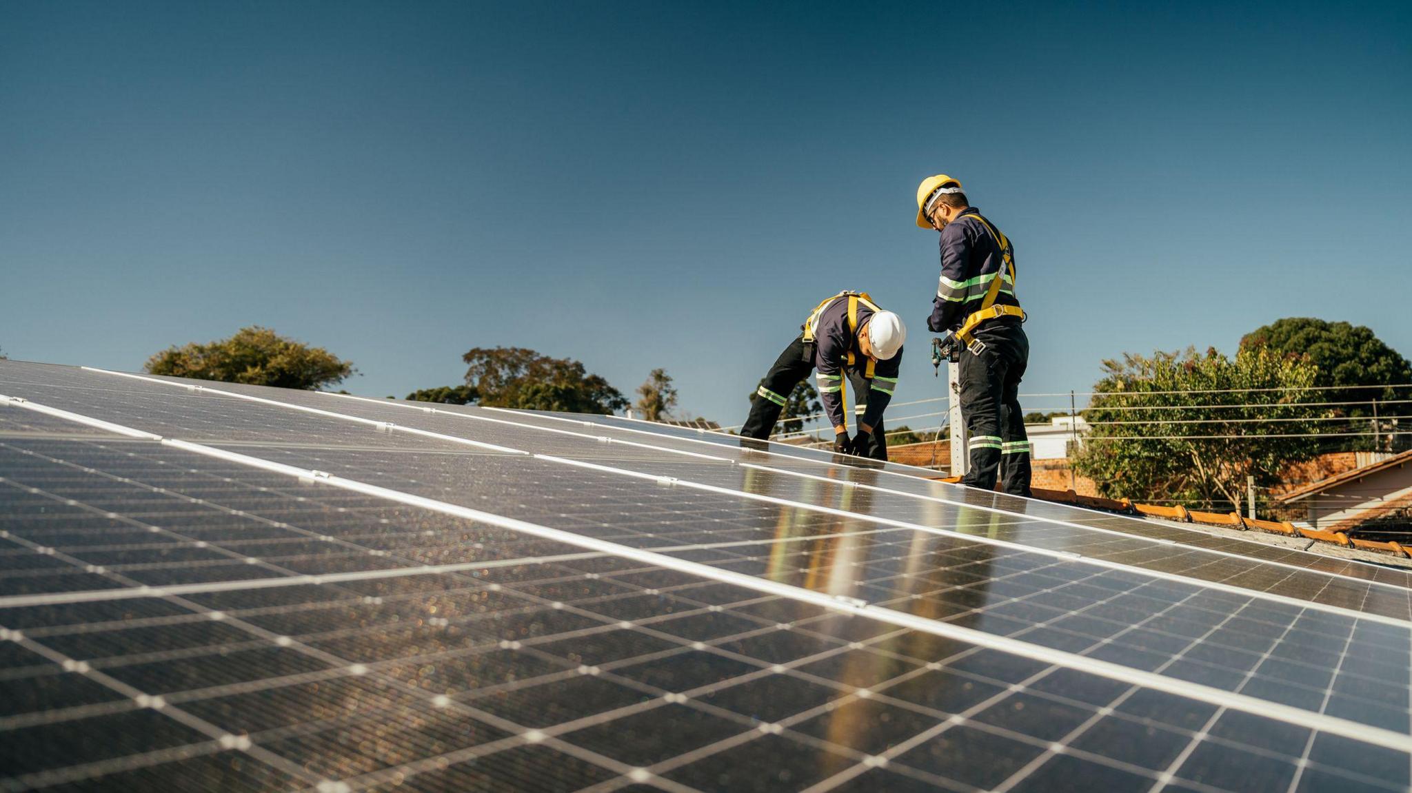 Two men in overalls and hard hats installing solar panels on a roof on a sunny day.