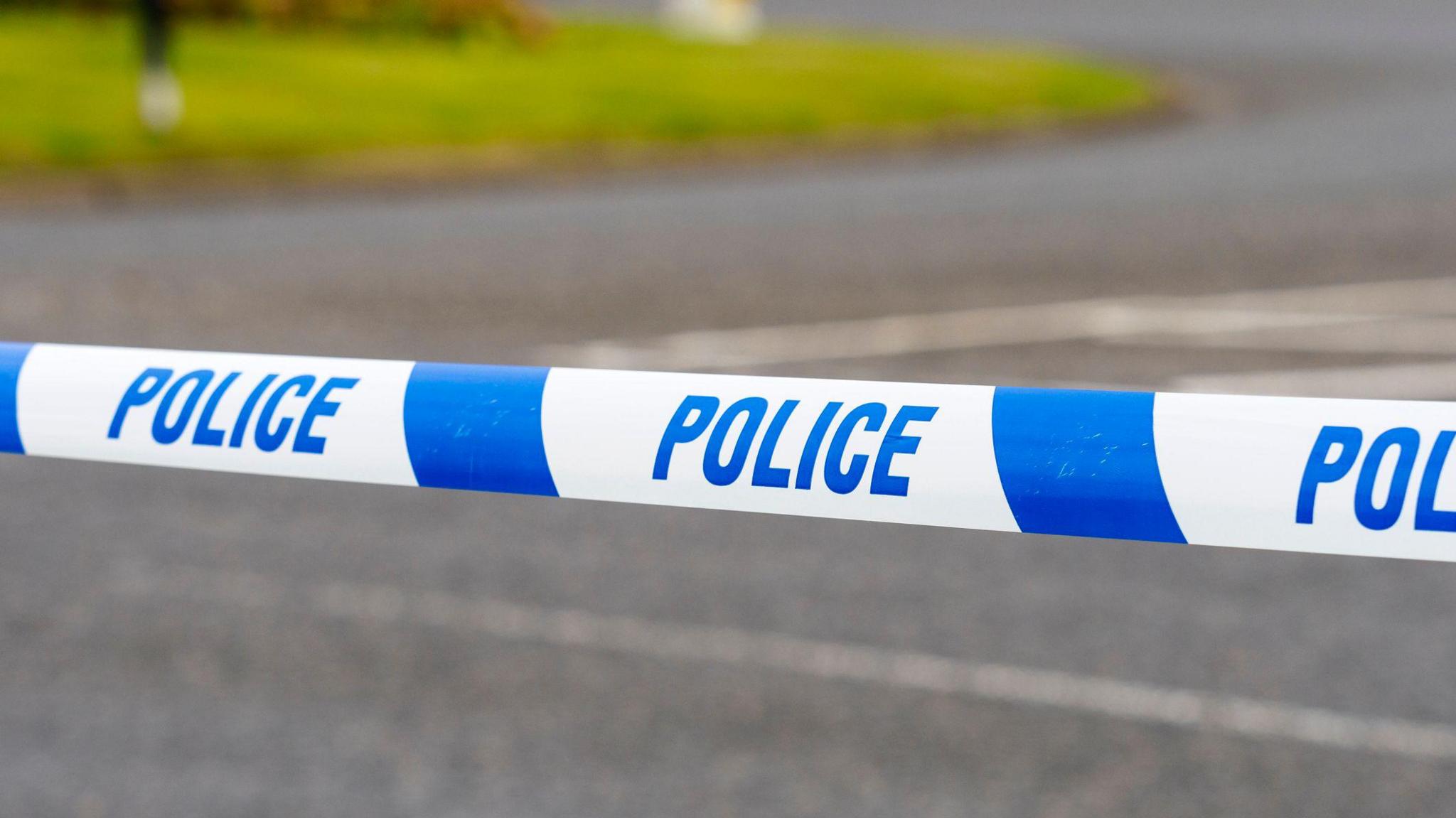 A line of police tape which is white and blue with the word police printed on it on repeat. In the background you can just see a concrete road and some grass