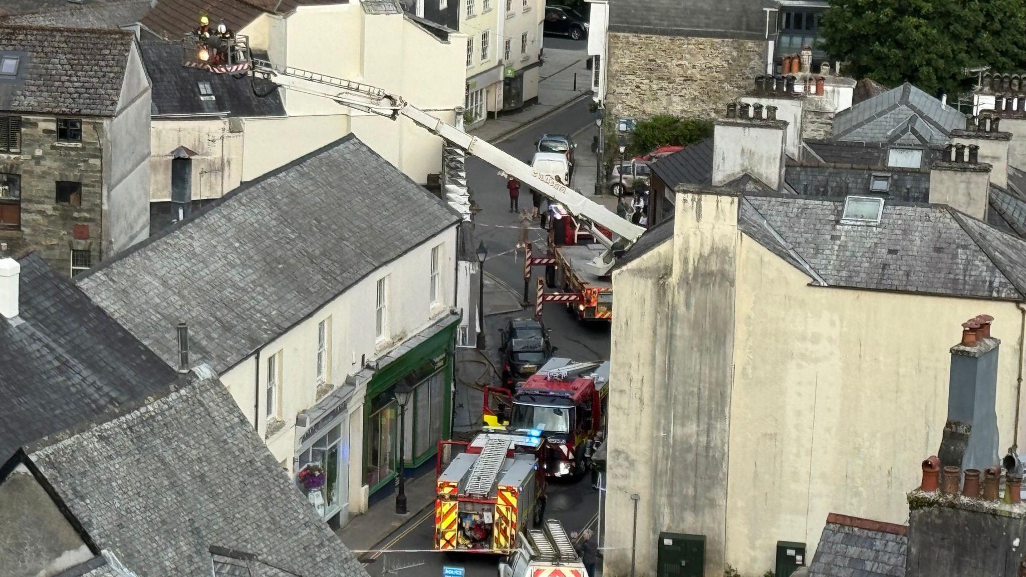 A hydraulic ladder with a team of fire fighters in tis cage is high over the rooftops of houses. There are four fire appliances on the streets below.