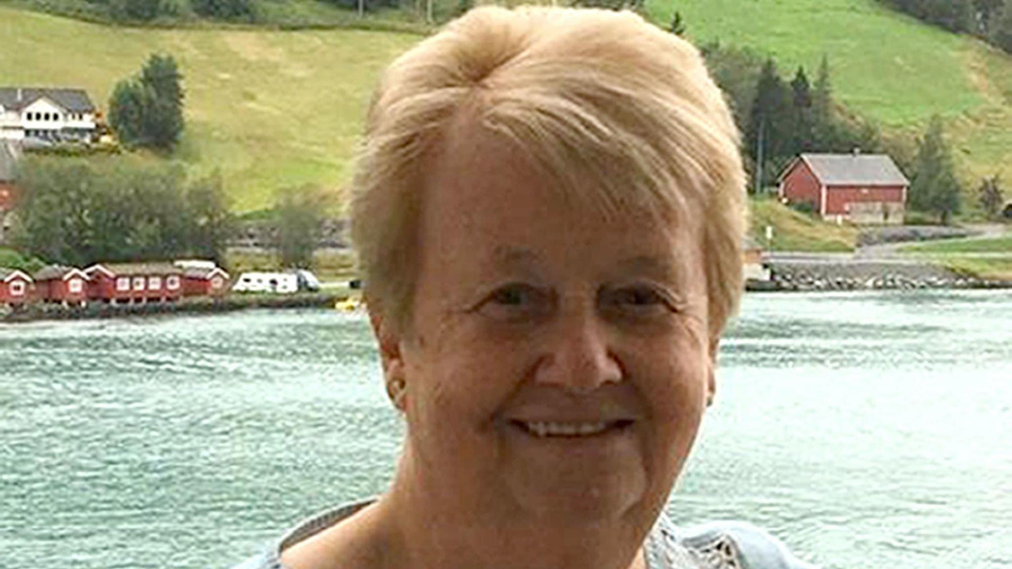 June Henty smiles at the camera, standing at a scenic location with an expanse of water behind her and grassy slopes, trees and a few houses further in the distance. She has neat, short, blonde hair and wears a light-coloured top.