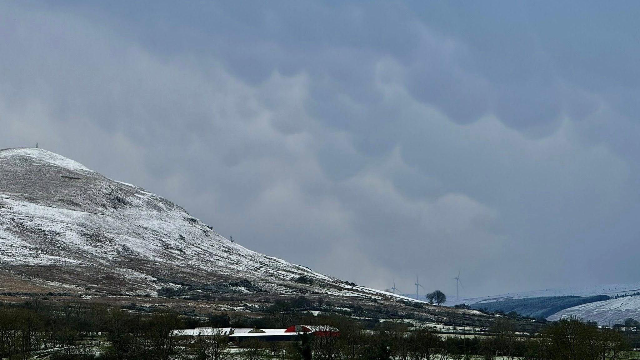A photograph of a winter landscape taken at the Sperrins on 20 November 2024. Snow and frost can be seen on the ground. A large hill also topped with snow is to the left of frame. A building can be seen among leafless trees below the hill. The sky is grey and cloudy and covers much of the frame. Three wind turbines can be seen faintly in the background. 