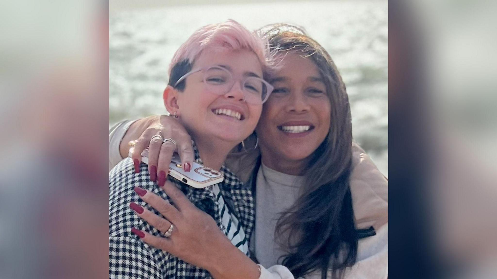 Too women smile at the camera, the one on the right has her arms wrapped around the other. The sea is behind them with the sun shining off it.