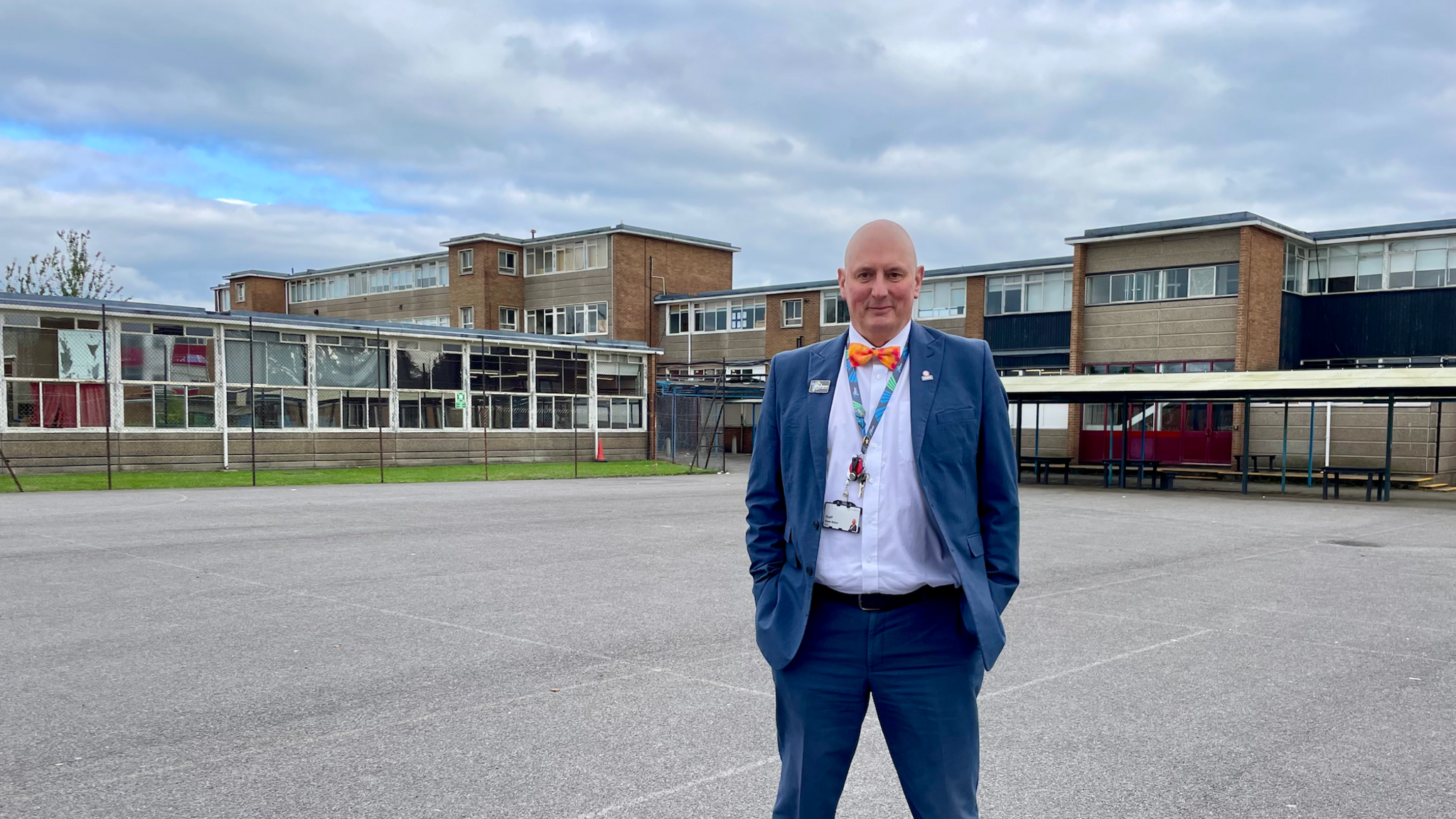 Dave Baker, wearing a blue suit, white shirt and orange bow-tie, stands in the playground at Patchway School, in front of some of the buildings that will be demolished