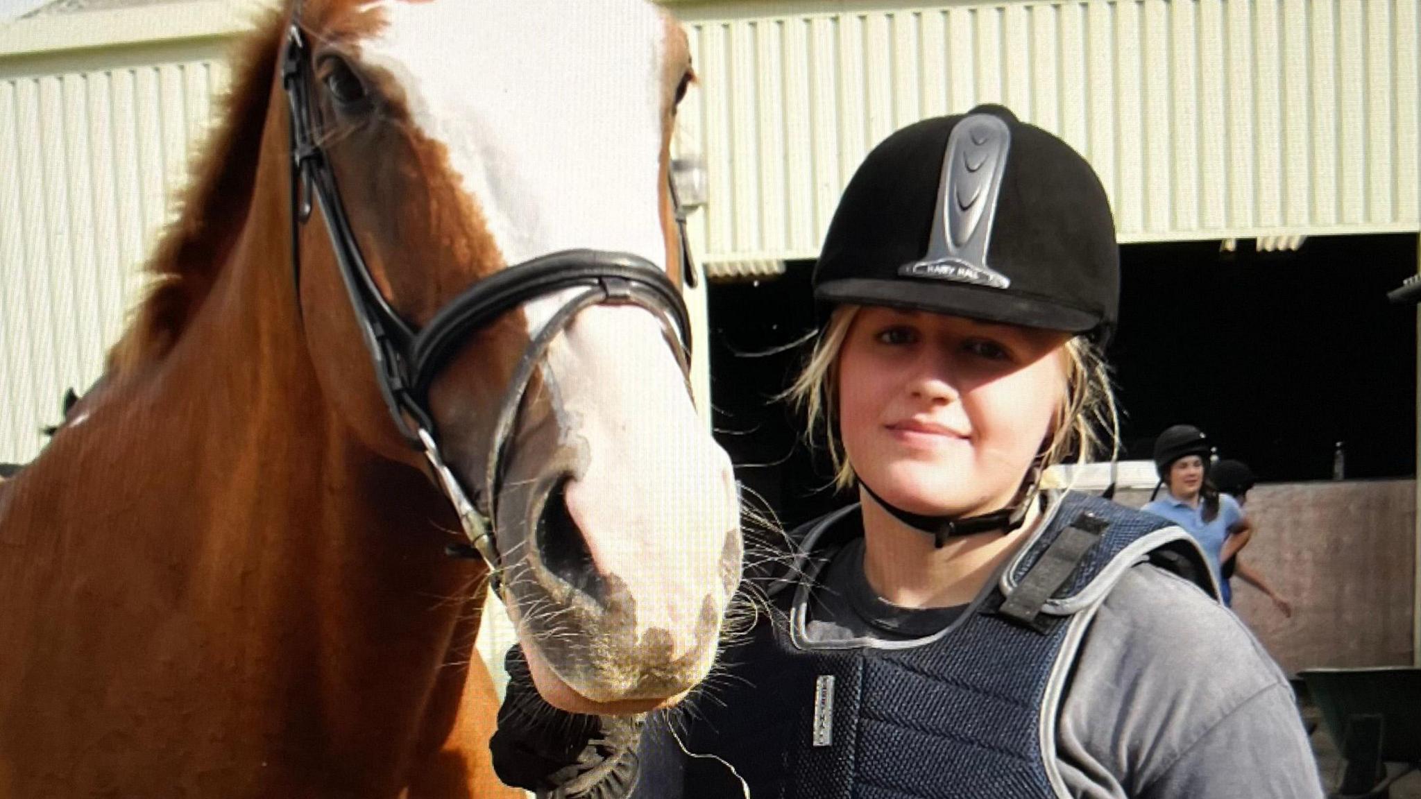 Abbi stands next to a horse at her riding stables. She is wearing a black riding helmet and has a chest vest on to protect her. She has strands of light blonde hair falling down around her face and a grey t shirt on. Abbi's horse has light brown with a white strip on its nose.