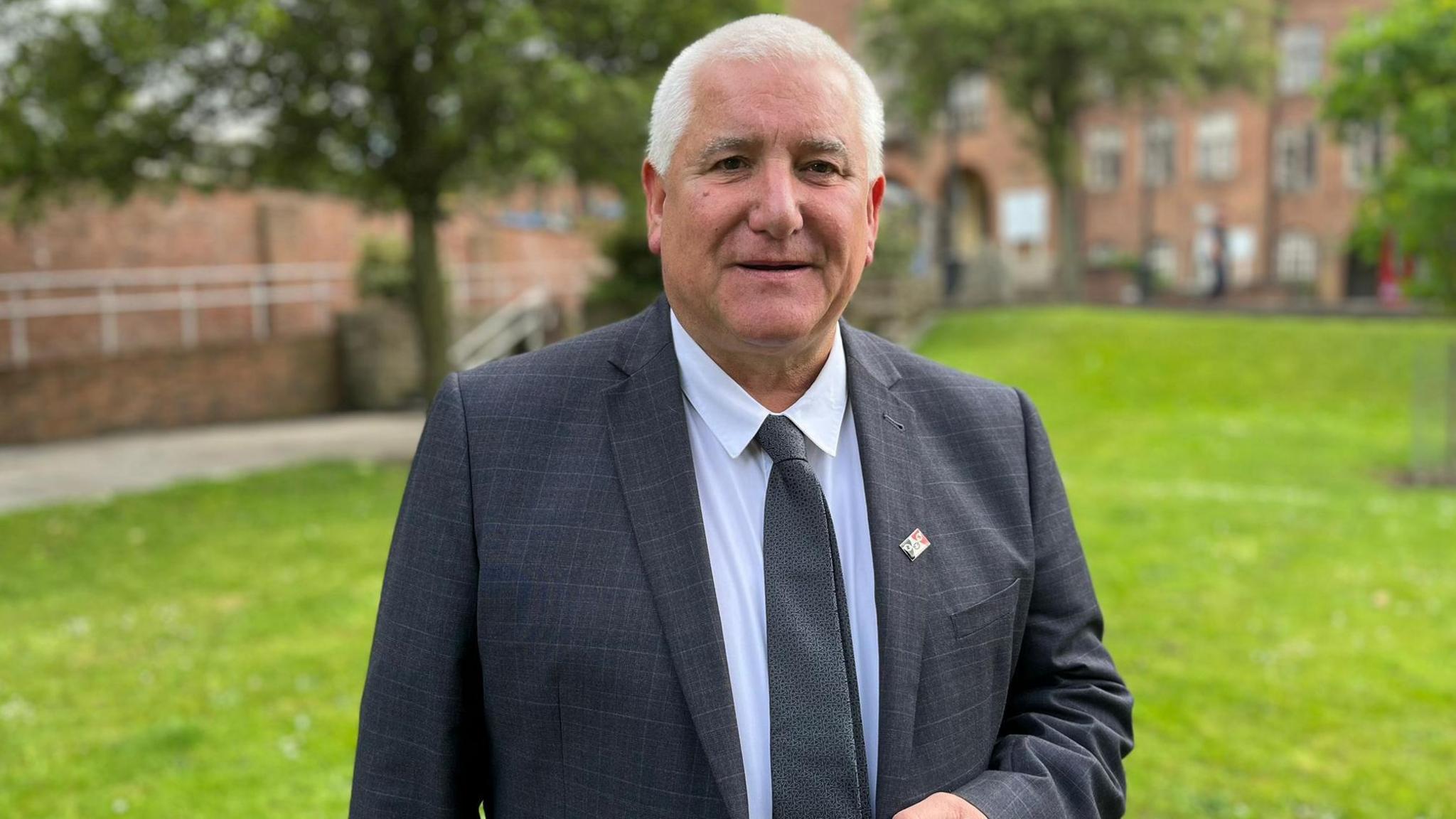 A man wearing a black suit and tie. He is wearing a small Black Country badge on his blazer. He has grey hair. In the background there is the Dudley council house.