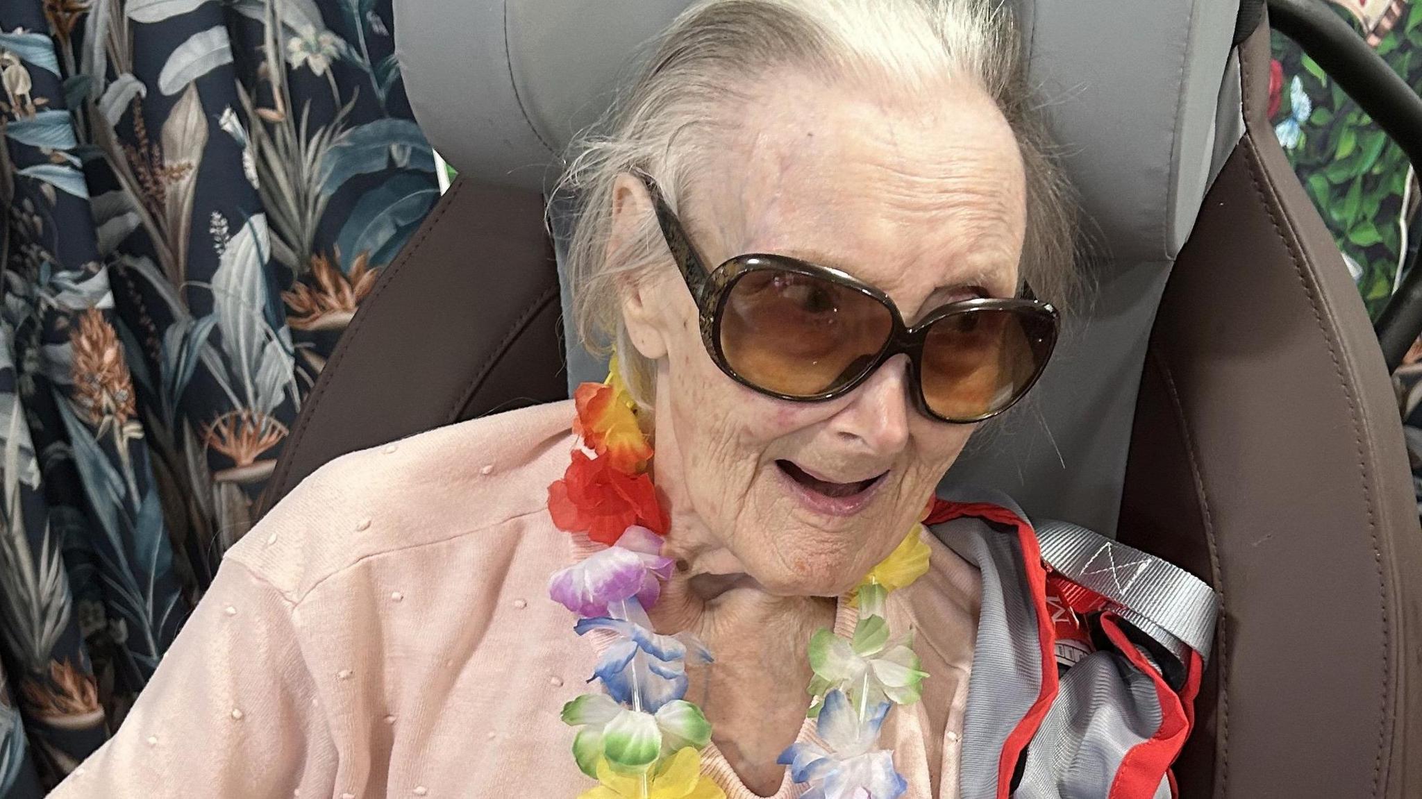 Joan sitting in a wheelchair with a pale pink jumper on, wearing dark sunglasses and a hawaiian flower garland smiling ar someone off camera