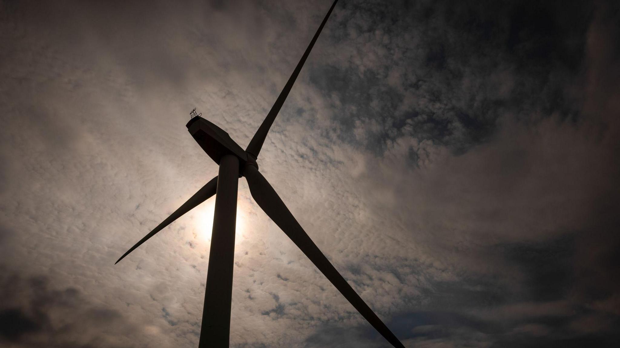 a wind turbine from below. It is silhouetted and against a blue sky with grey clouds. the sun is shining behind it