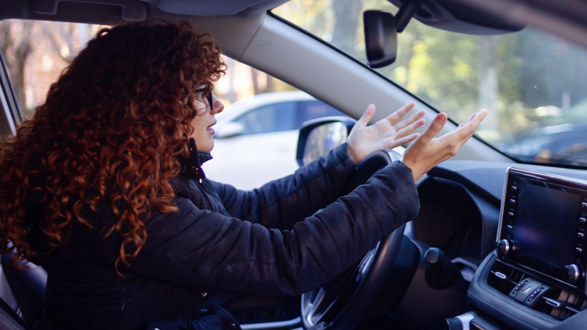 A woman with long red curly hair is sitting behind the wheel of her car, throwing her hands up in the air in frustration. She is wearing glasses and a black coat. The interior of the car is black and you can see trees, out of focus, beyond the windscreen.