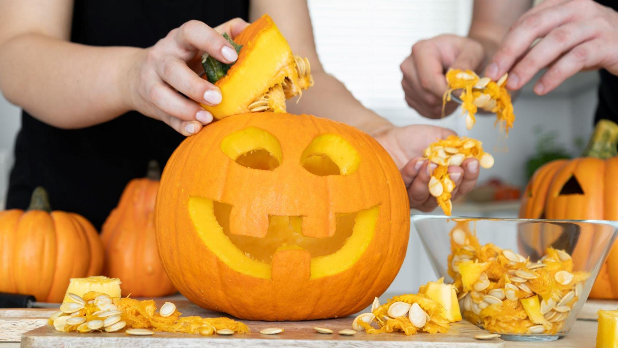 A person pictured from the shoulders down is holding the top of a carved pumpkin in one hand, and in the other is holding pumpkin seeds which have been carved out. The pumpkin is sitting on a table beside a glass bowl which is full of pumpkin seeds. A person's hands are pictured on the right holding a spoon full of pumpkin seeds.