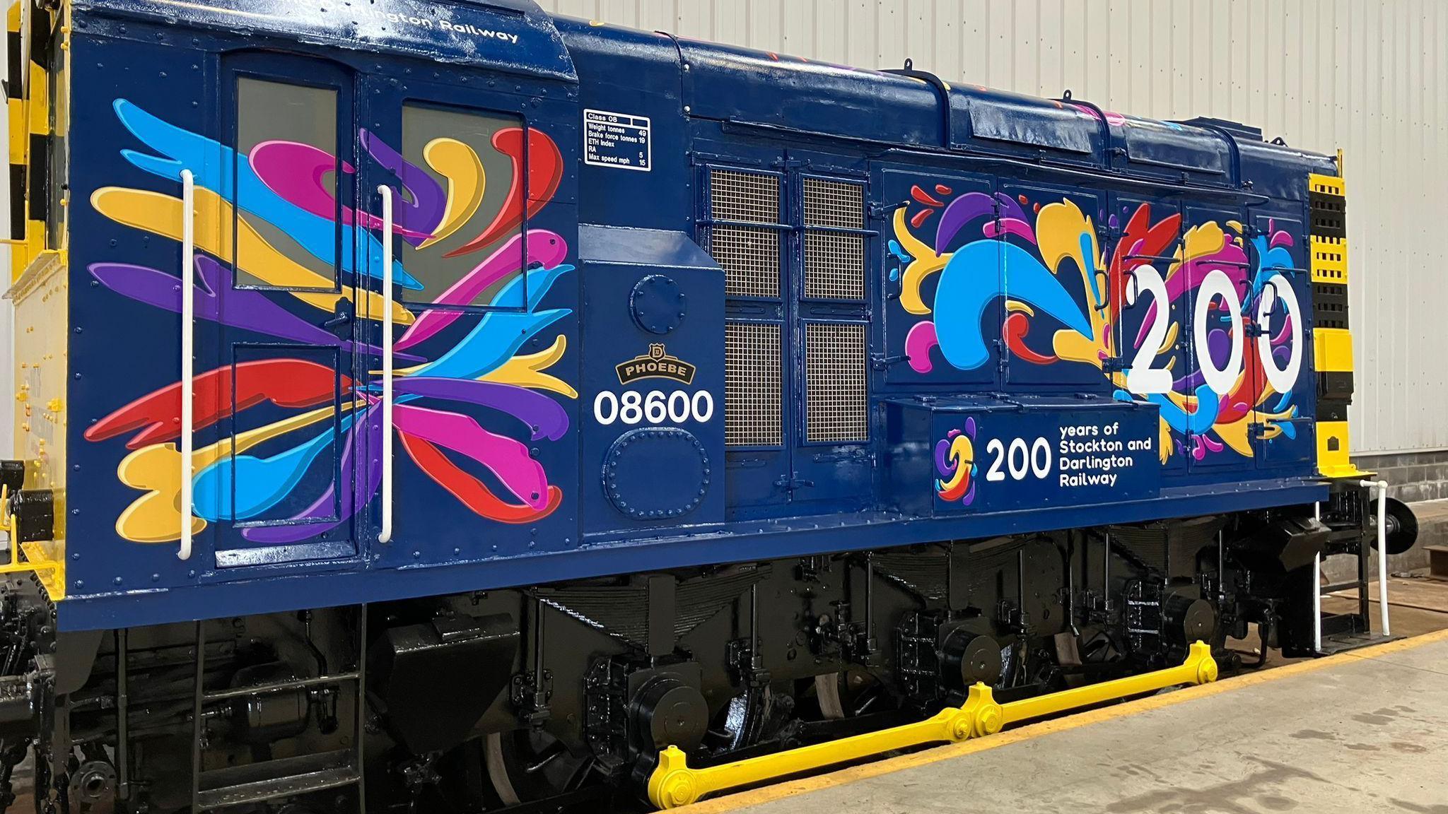 A dark blue shunting engine with colourful splashes of paint and a big "200" in white letters, beneath which it reads "years of Stockton and Darlington Railway". The engine is rectangular shaped and the four wheels beneath it are shining black with bright yellow rods connecting them.