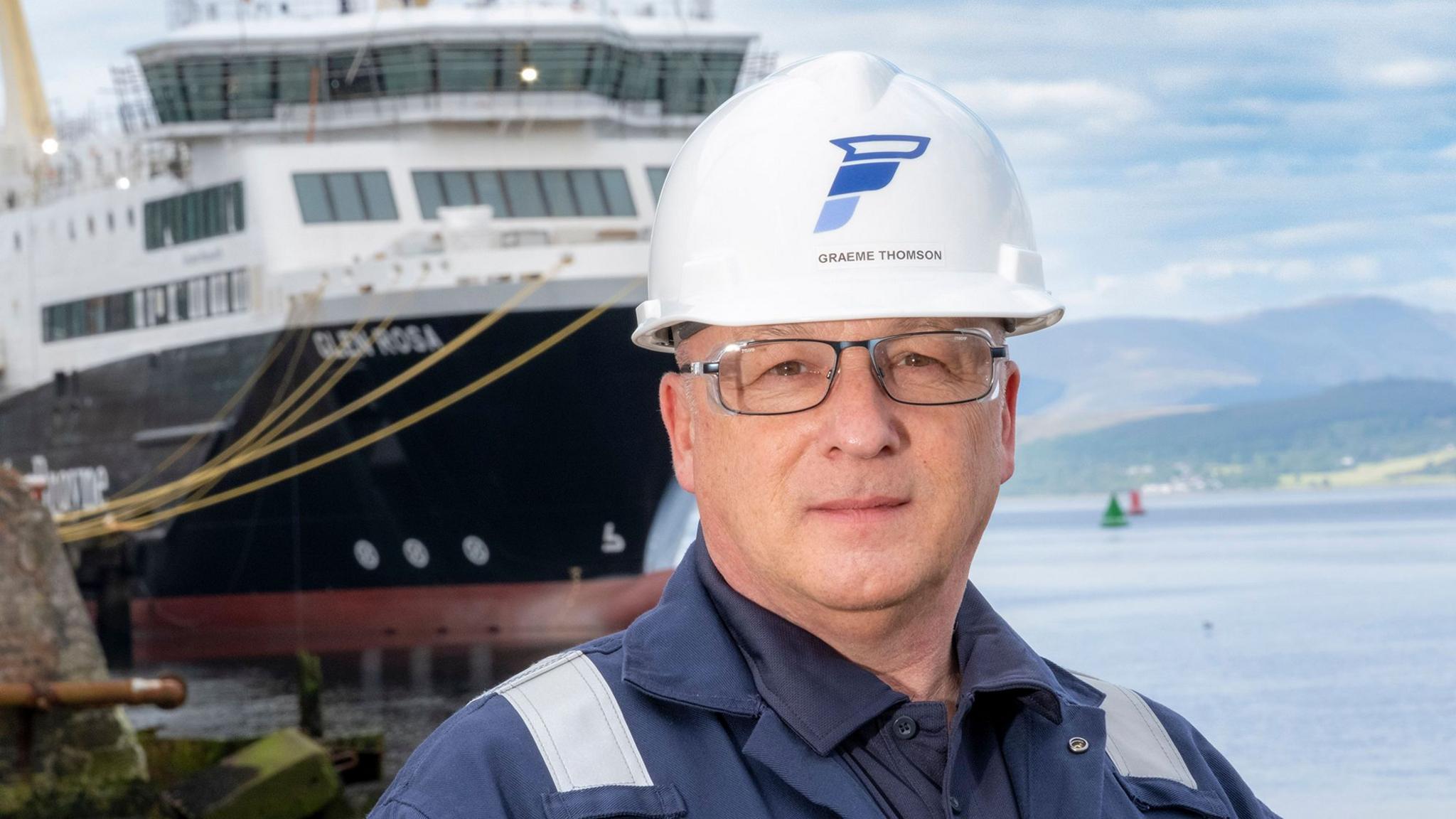 A bespectacled man in blue overalls and a white hard hat. The large ferry Glen Rosa can be seen in the background