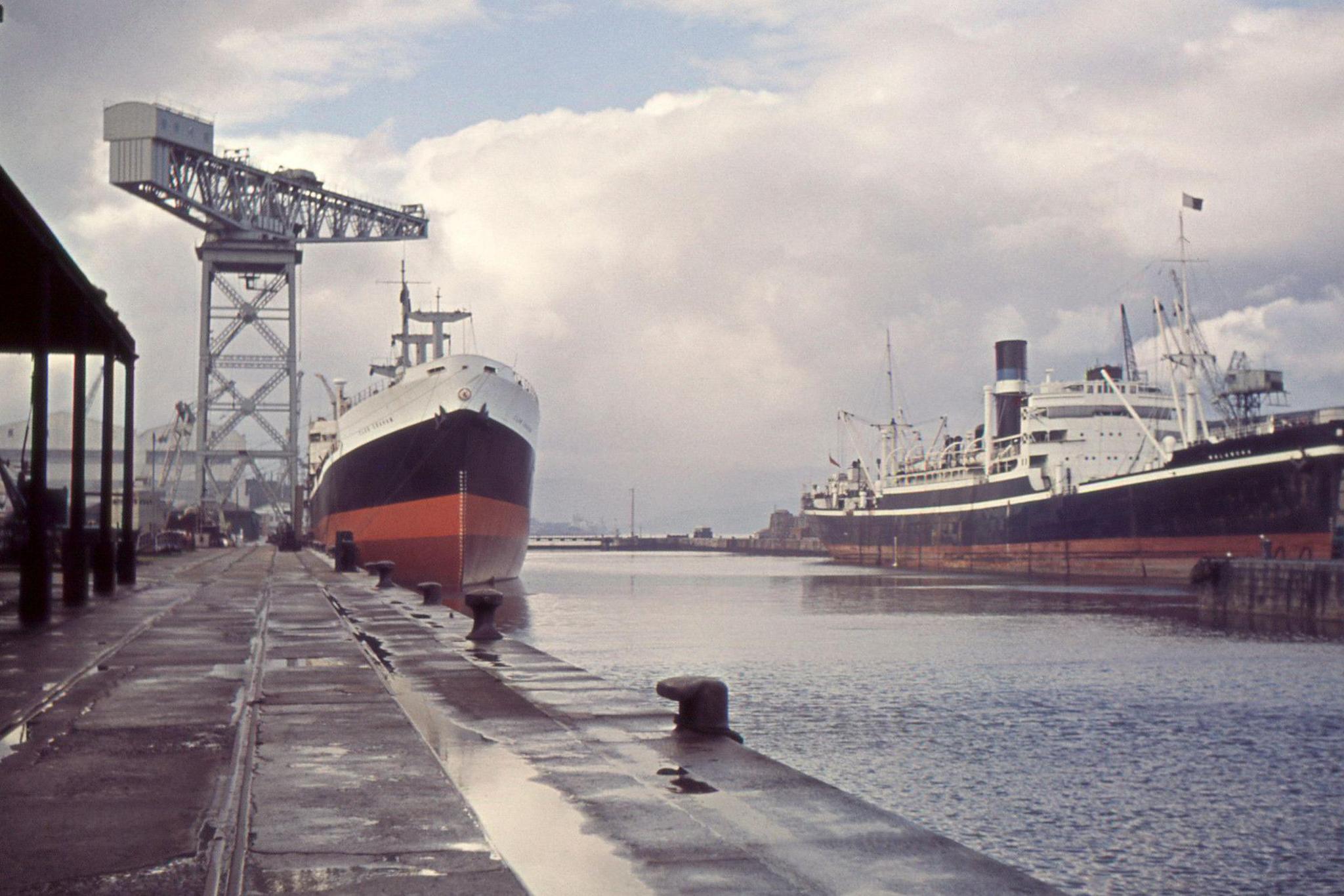 A crane stands in the distance next to a black red and white ship which is moored in a dock. Another ship is moored on the other side of the dock