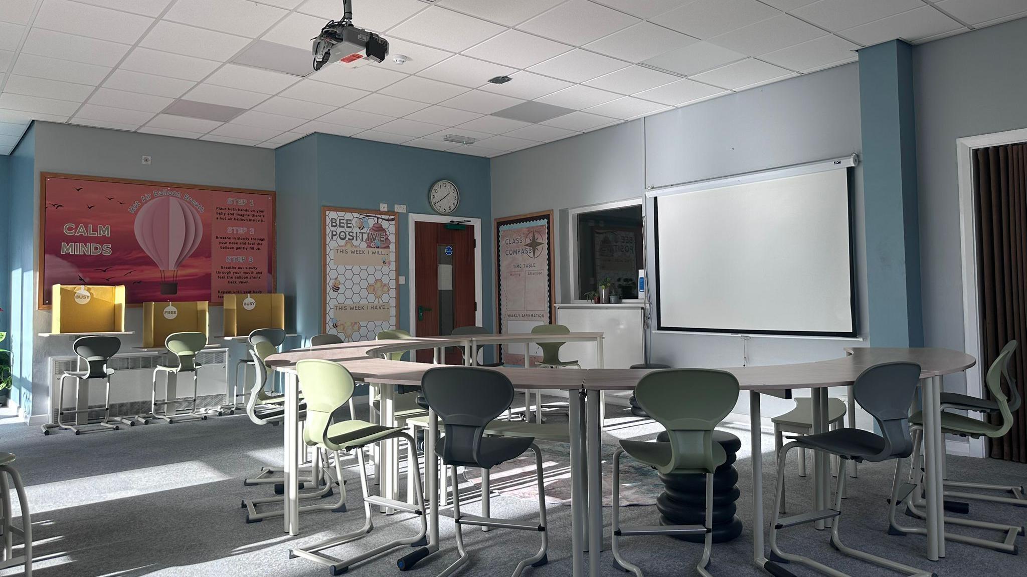 The adaptive classroom from behind, you can see arched tables, there are ergonomic green chairs facing a projector.