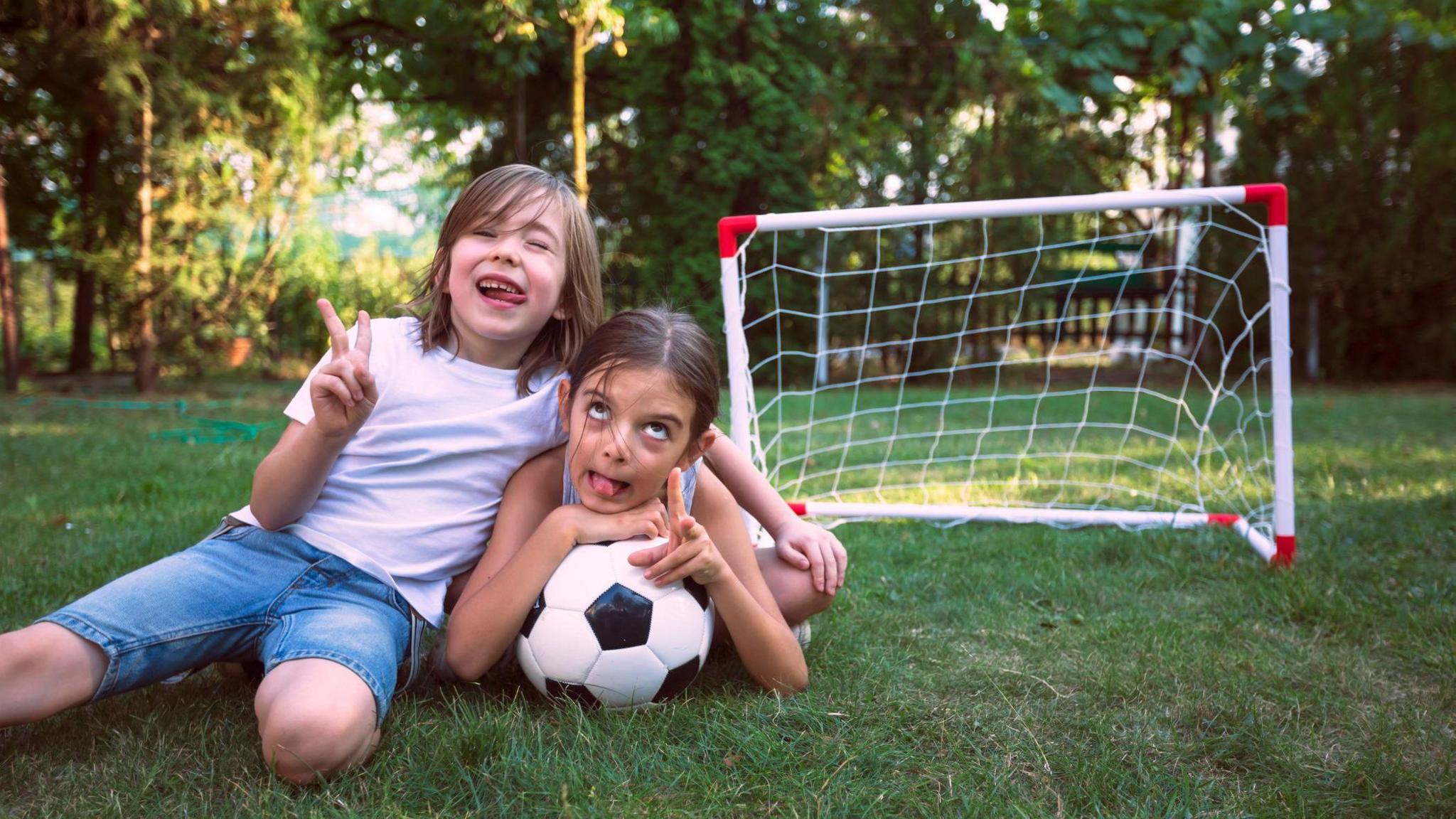 Two children pulling funny faces while on the ground with a football. 