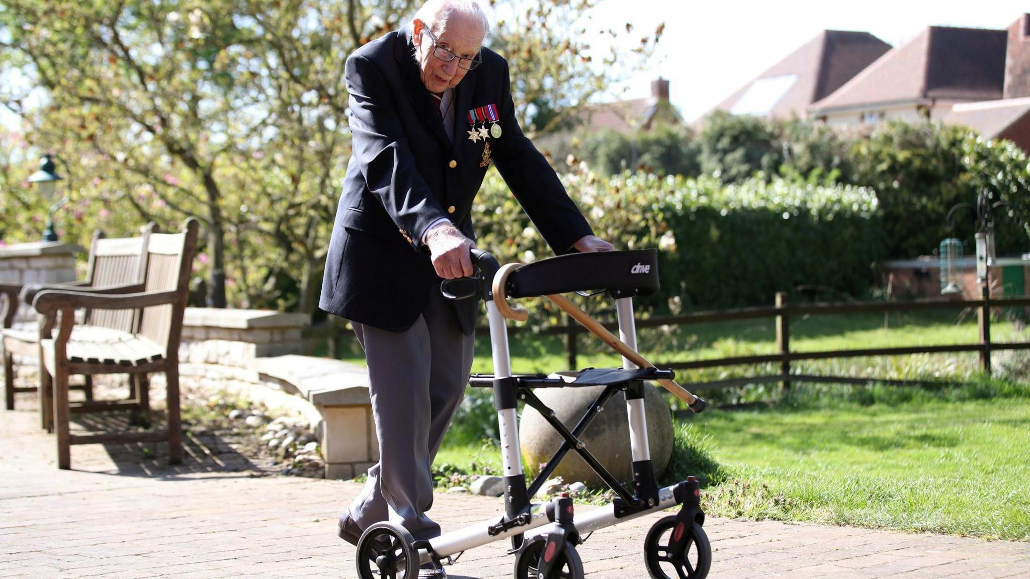 Captain Sir Tom Moore at the age of 99, walking around his garden to raise money for the NHS during the pandemic. He is holding a sturdy zimmer frame on wheels and wears grey trousers and a dark blue blazer, with his medals on his left pocket. He has glasses on and is hunched slightly and glancing sideways at the camera. In the background are lawns and houses, and a low wall and bench behind him.