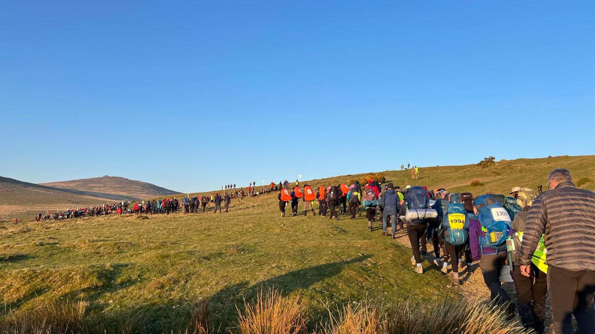 Masses of young people are walking a trail that winds up the top of a tor on Dartmoor. The terrain has large tufts of grass and pieces of granite scattered across it.