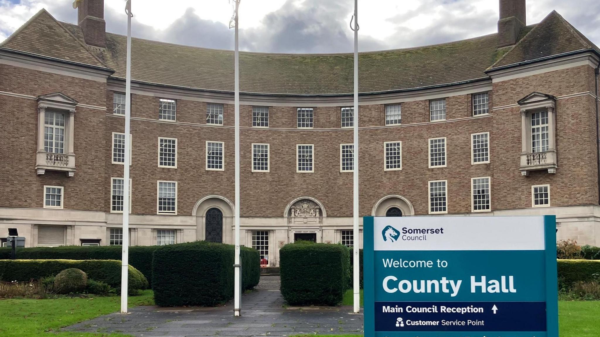 A sign reading "welcome to county hall" outside a large municipal building with flags flying on a sunny day